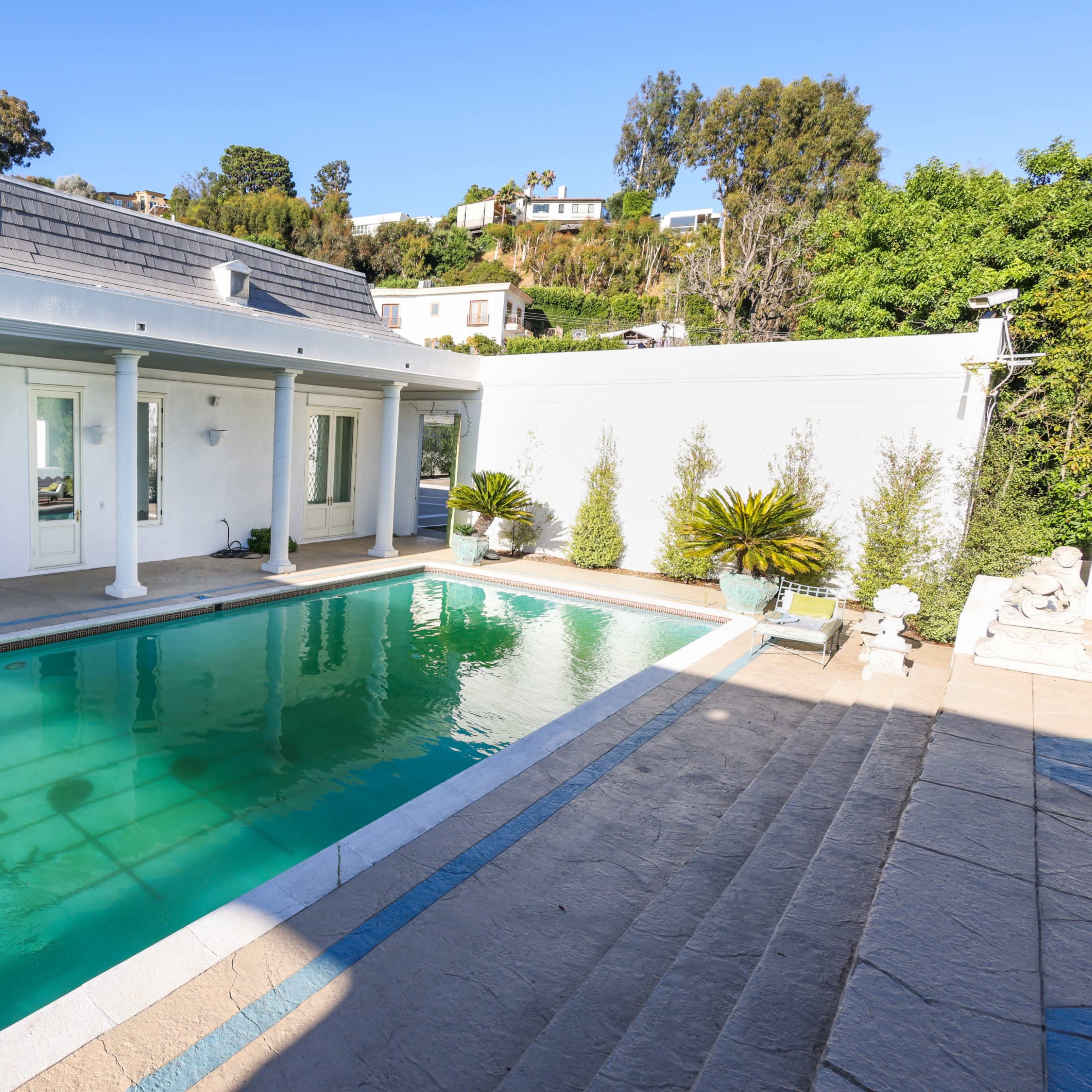 A modern swimming pool is surrounded by a white house with large windows and greenery in the background.