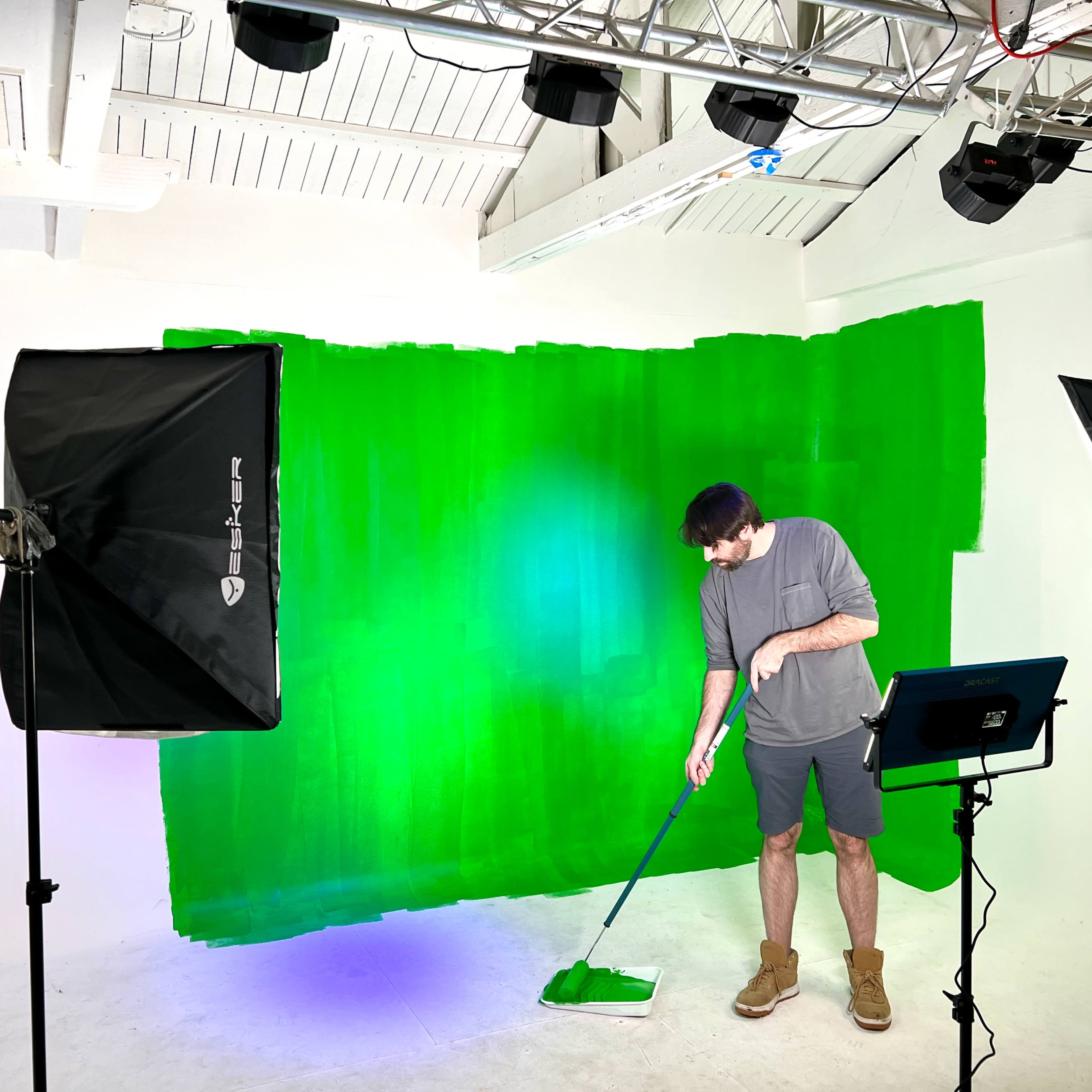A person is painting a green backdrop in a photography studio while standing next to lighting equipment and a tablet.