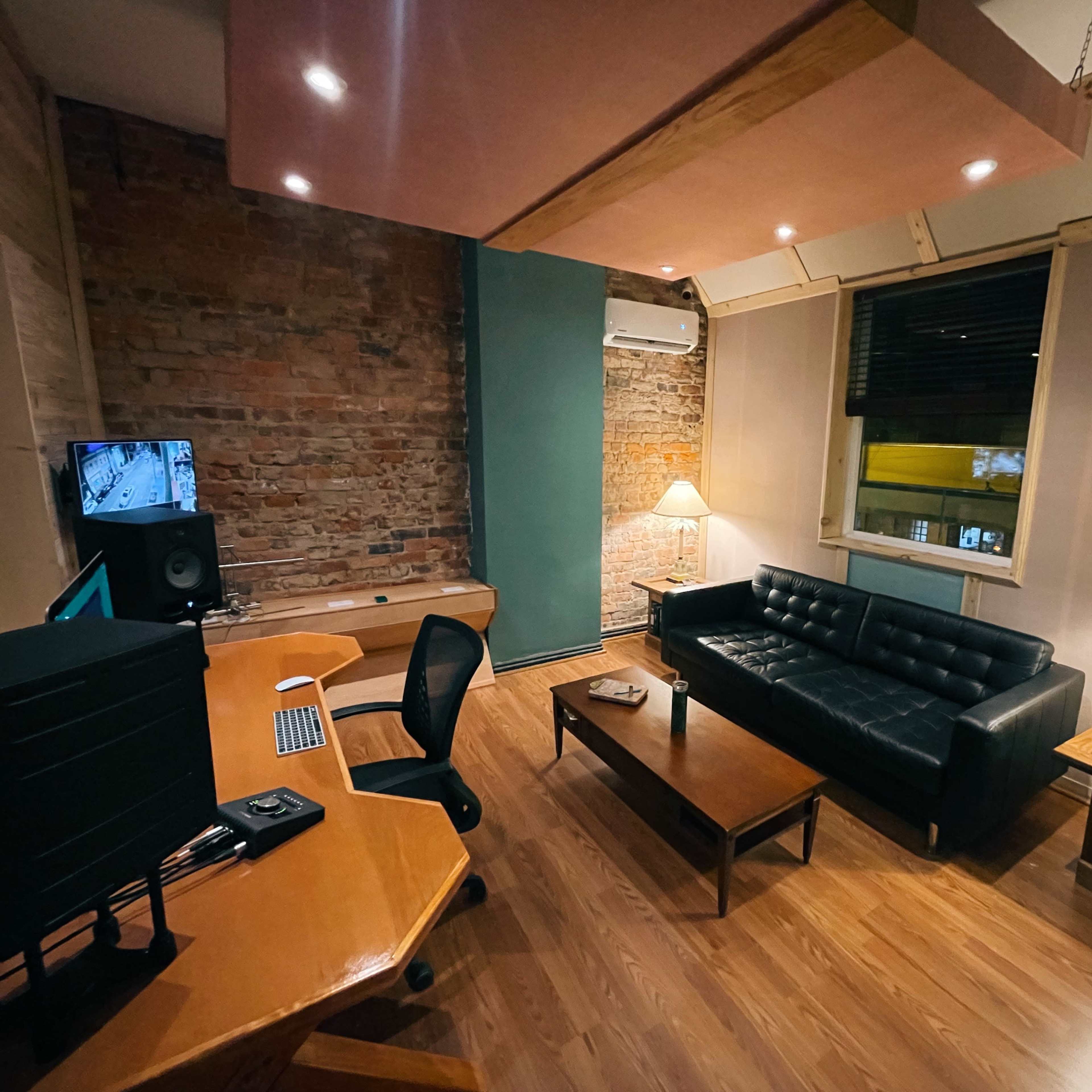 A cozy room with a black couch, a wooden coffee table, an office desk with a computer, and two lamps, featuring exposed brick walls and a large window.