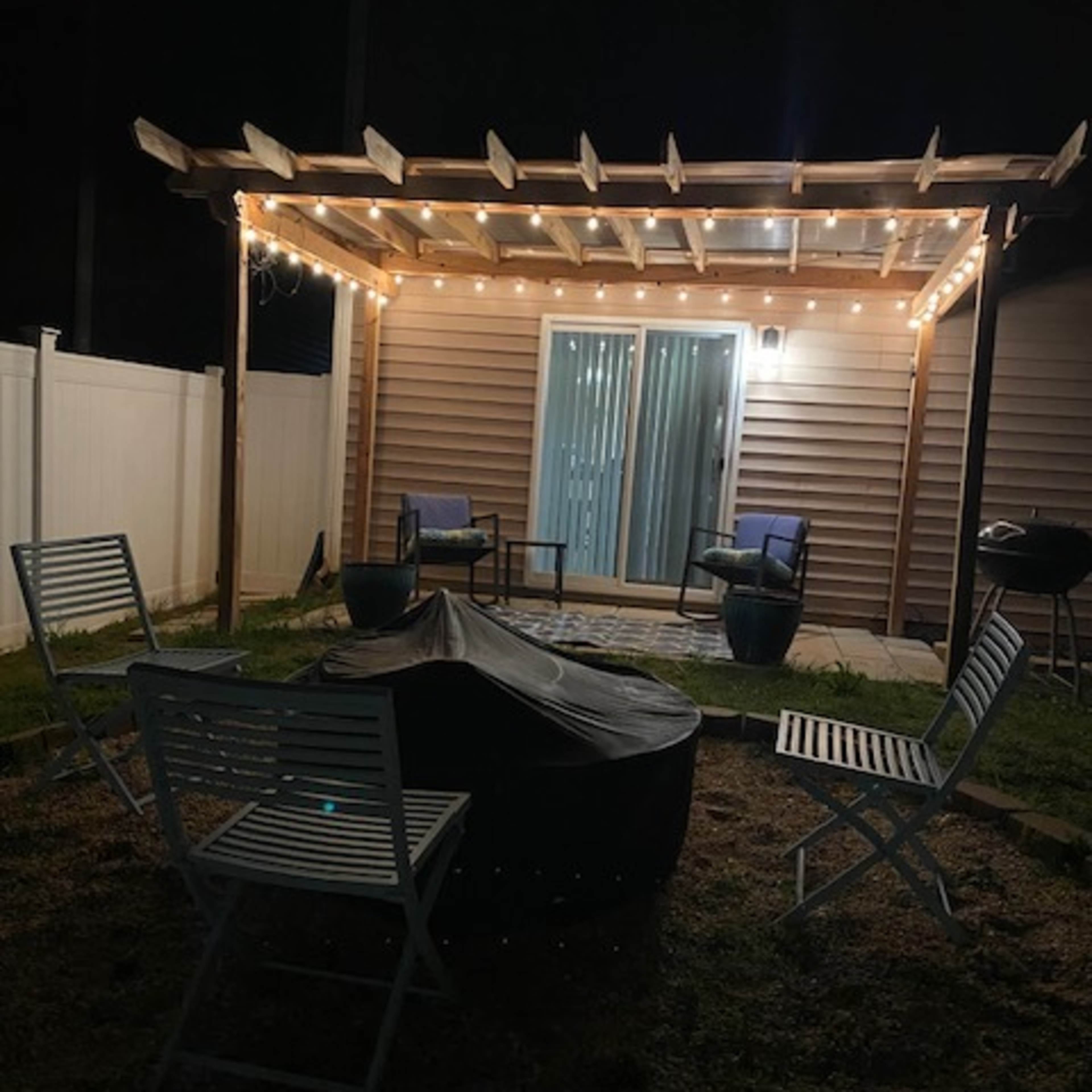 A patio area at night features a pergola with string lights, two chairs, and a circular fire pit surrounded by metal folding chairs.