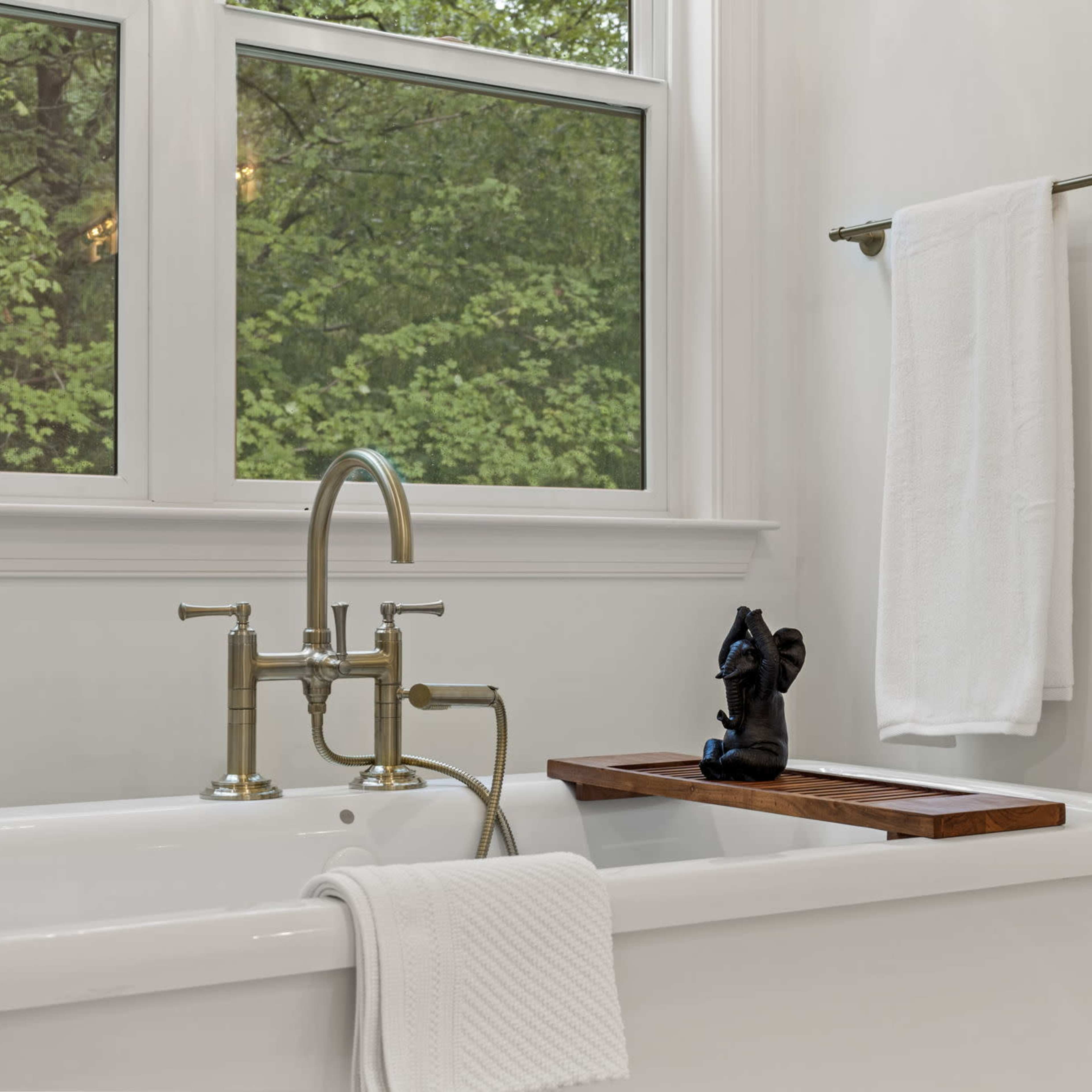 A modern bathroom features a freestanding bathtub with a wooden tray, two white bath towels, and a window overlooking greenery.