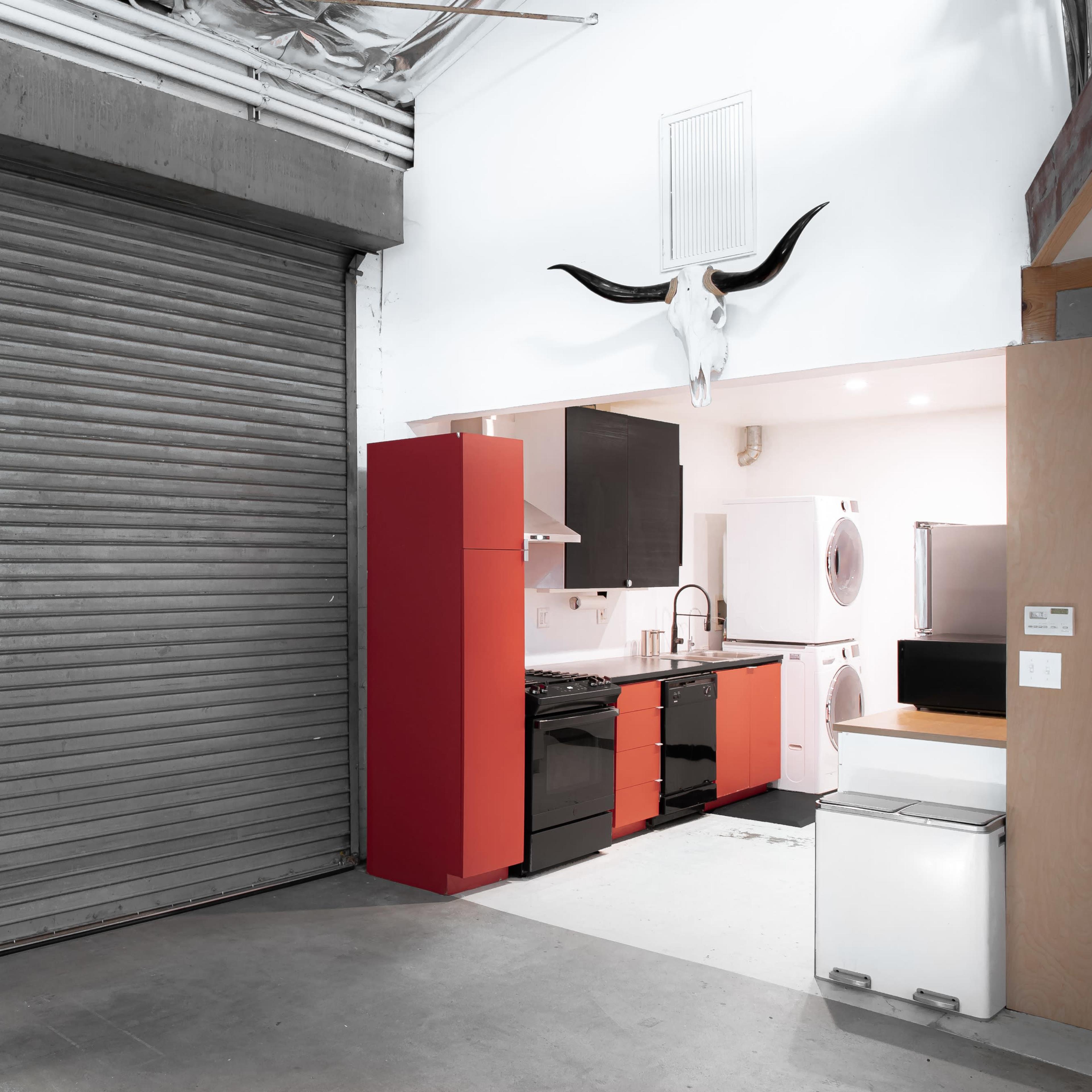A modern kitchenette with red and black cabinets adjacent to a large metal roll-up door in a spacious industrial-style room.