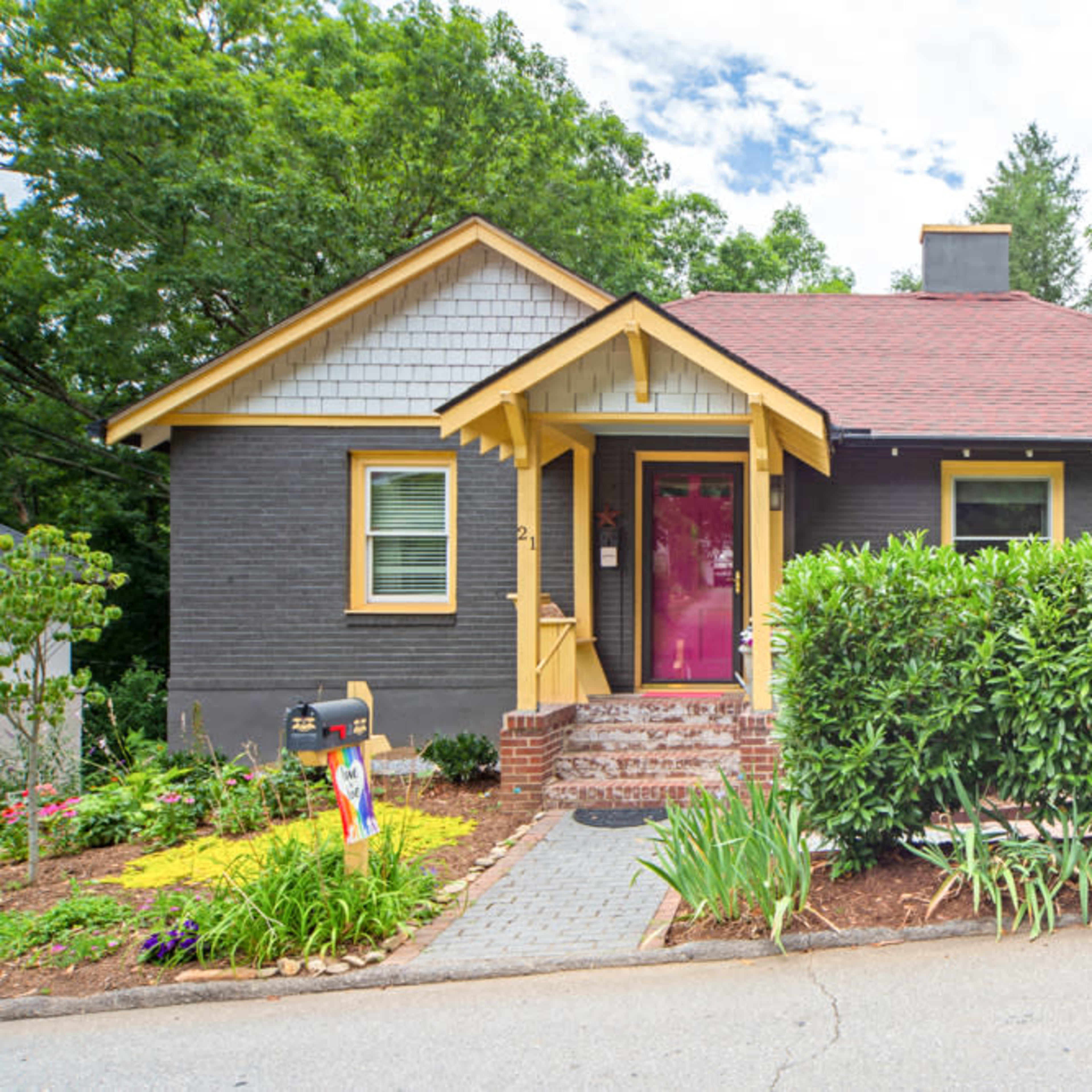 A one-story gray house with a red roof and a bright pink door is surrounded by green shrubs and a landscaped front yard.