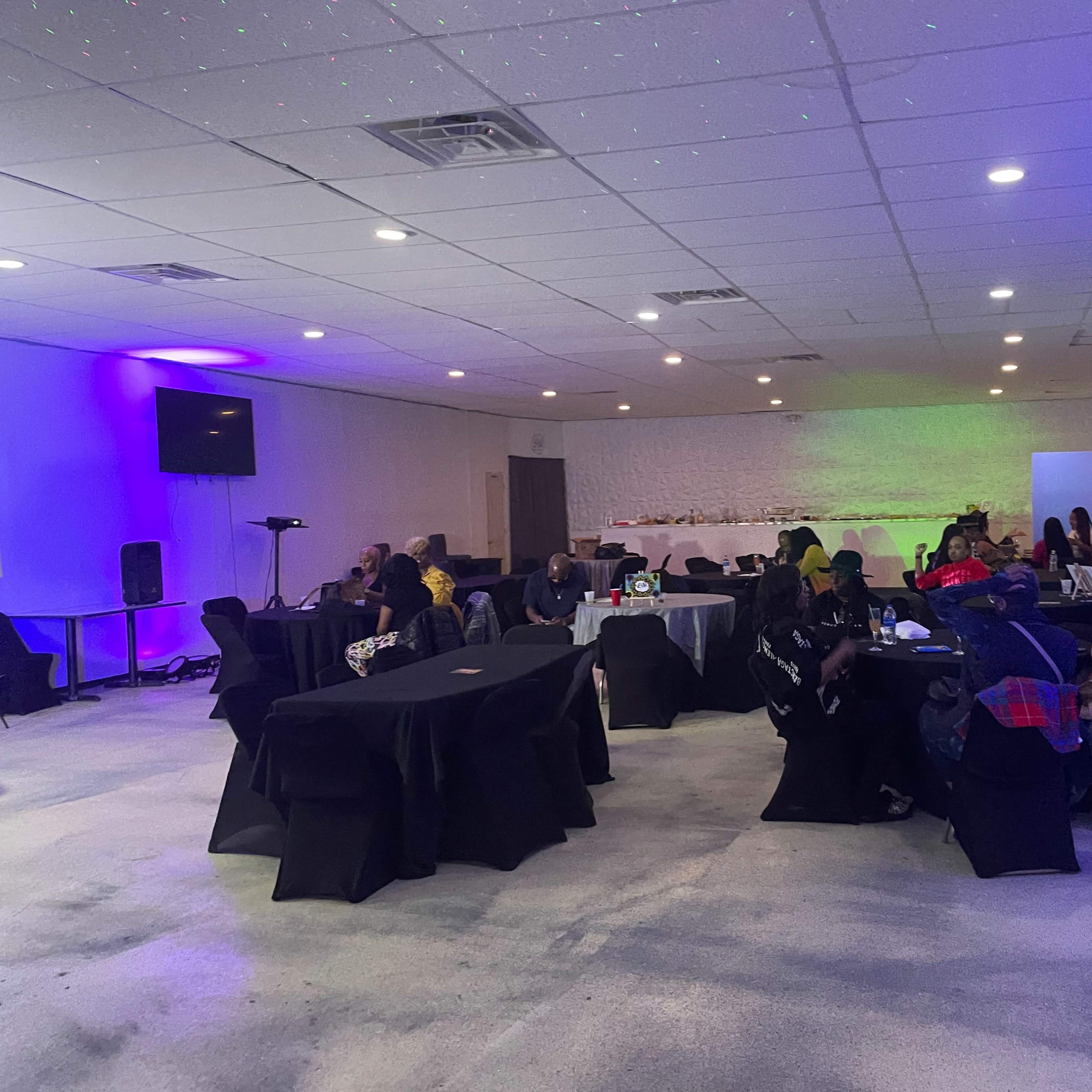 A group of people sits at tables in a dimly lit room with purple lighting and a projected logo on the wall.