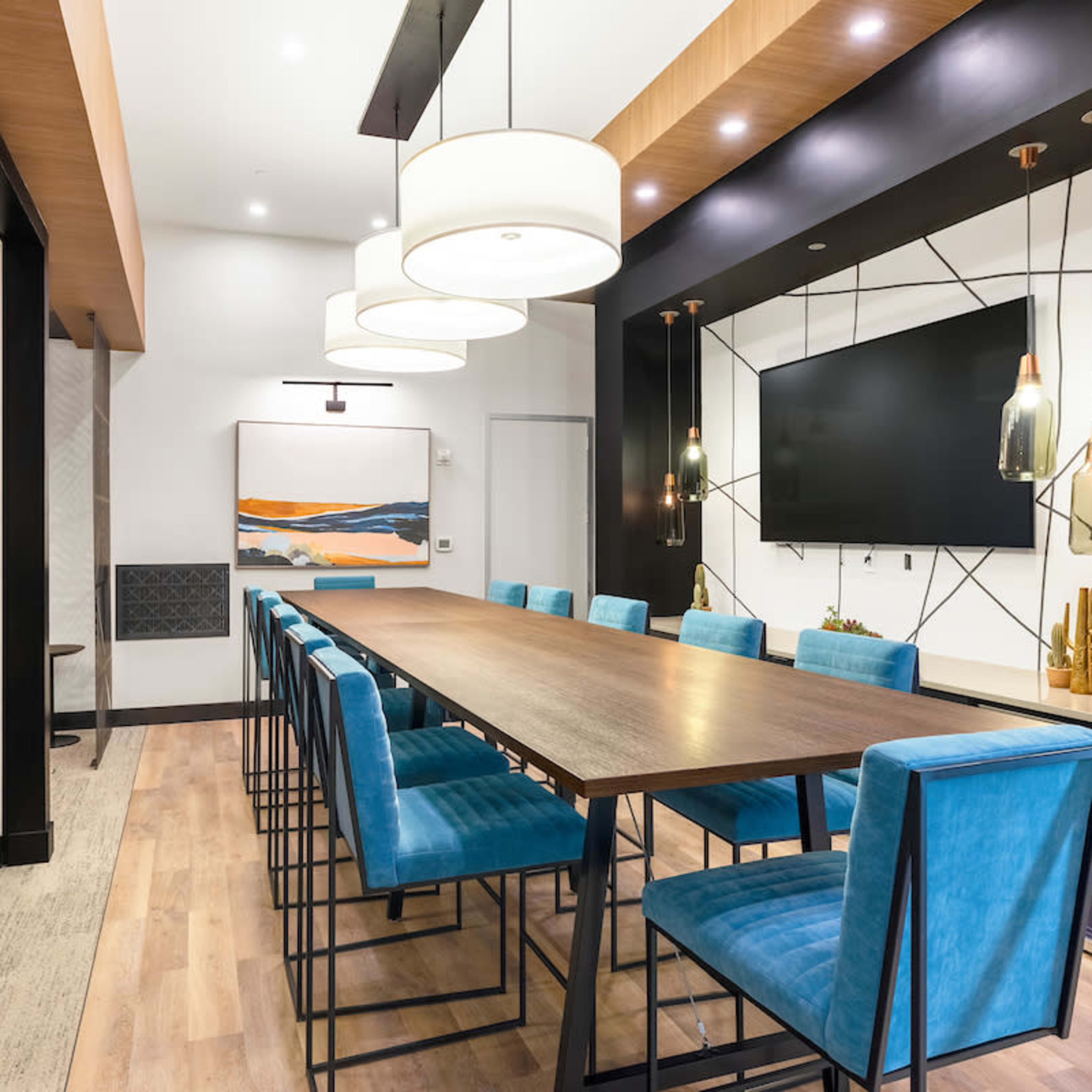 A long wooden table is surrounded by blue upholstered chairs in a well-lit dining area featuring a television and decorative wall art.