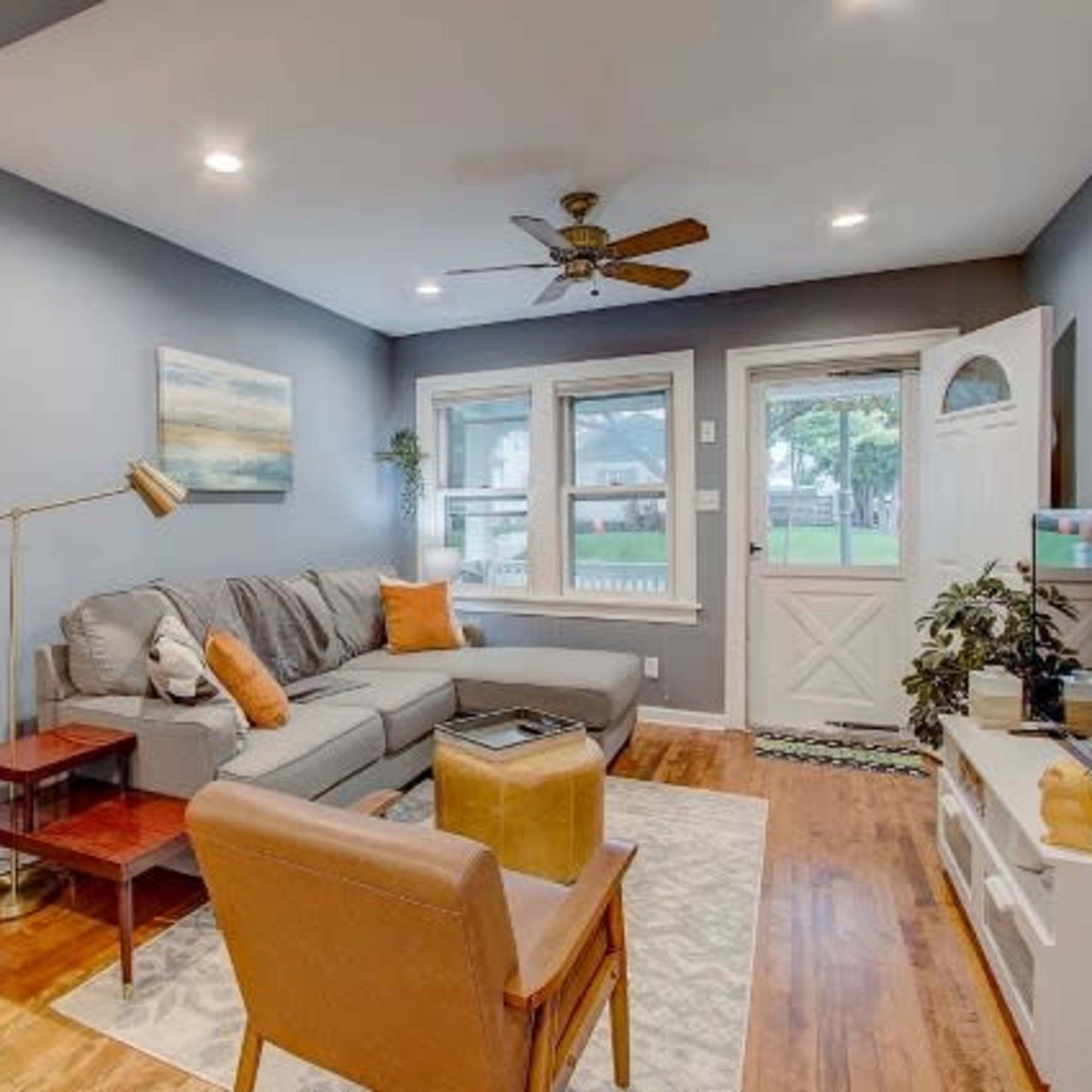 A living room with a gray sectional sofa, a wooden side table, an armchair, and large windows leading to an outdoor area.