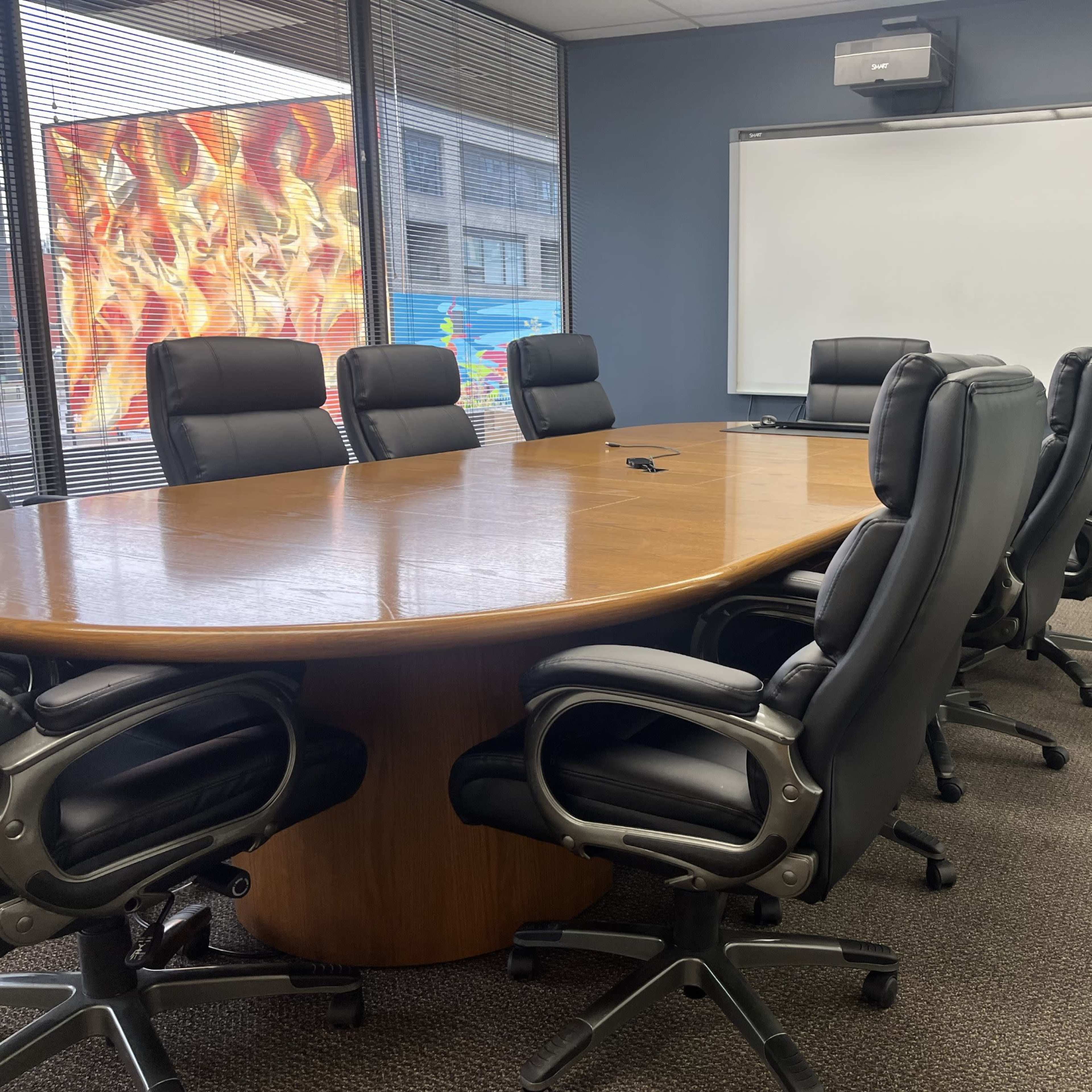 A large conference room features a circular wooden table surrounded by black leather office chairs, with a whiteboard and projector mounted on the wall.