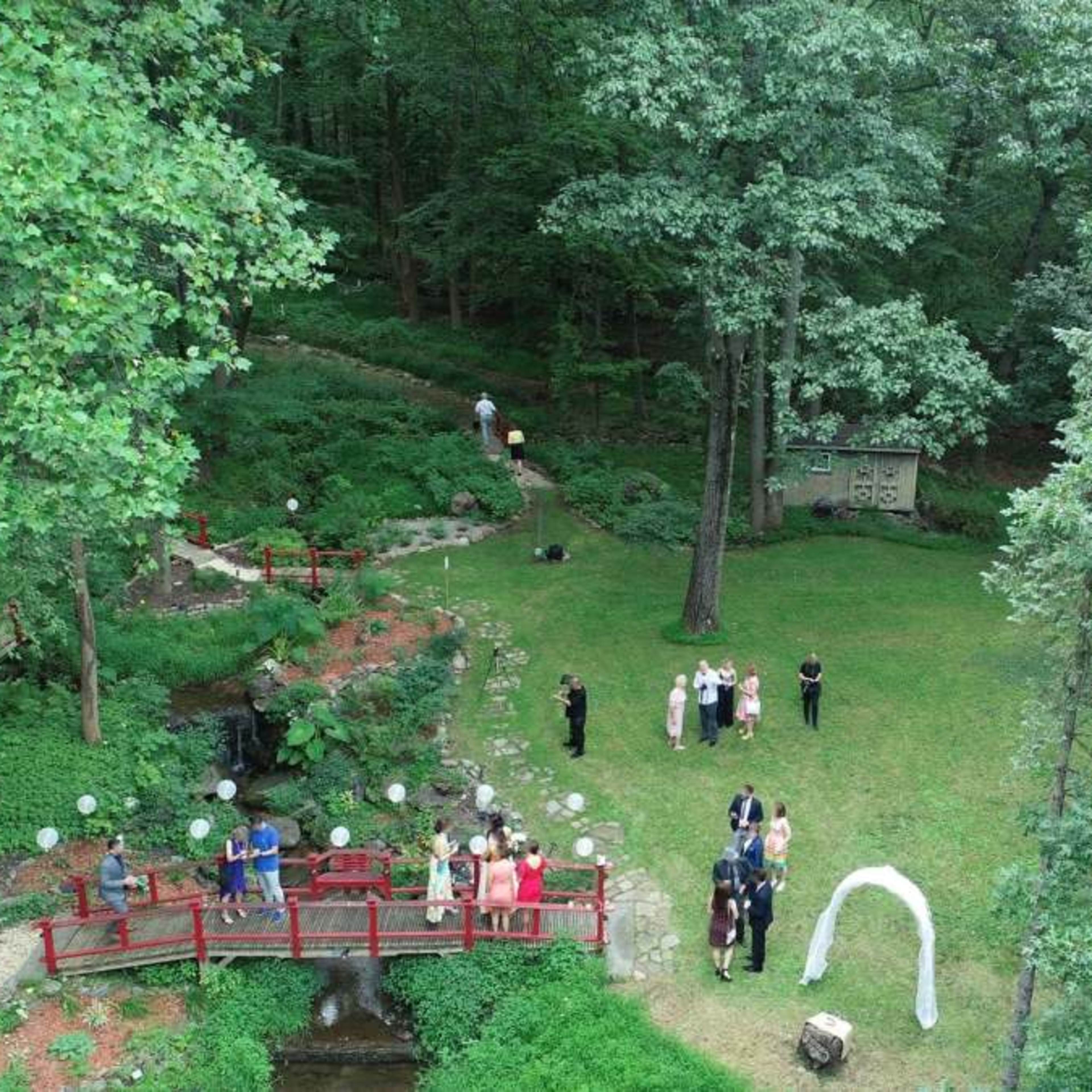 A wedding ceremony is taking place in a green garden, featuring a red bridge, a white arch, and guests gathered on the lawn.