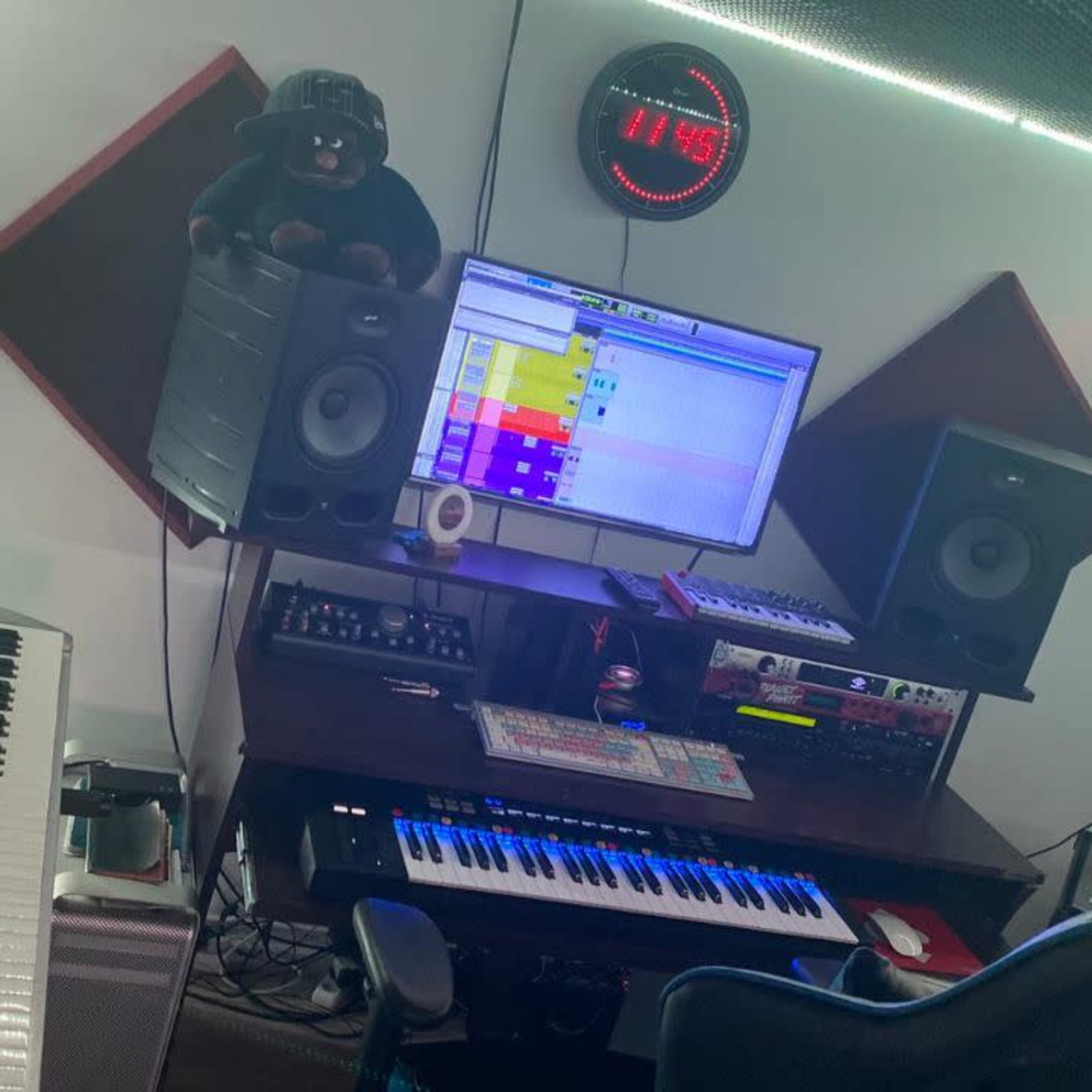 A music production workspace featuring a keyboard, speakers, a computer monitor with software open, and acoustic panels on the walls.