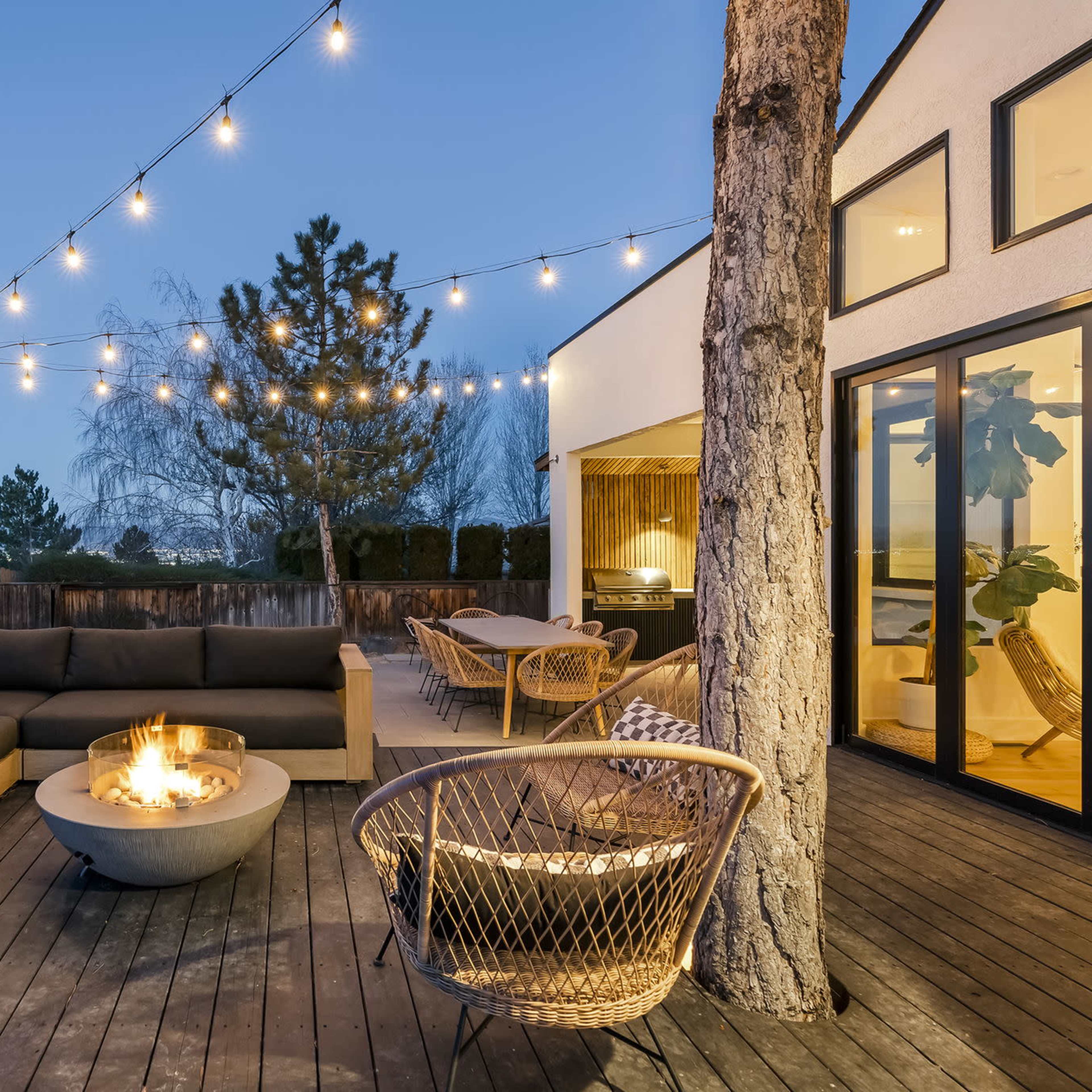 A modern outdoor living space features a sectional sofa surrounding a fire pit, illuminated by string lights at dusk.