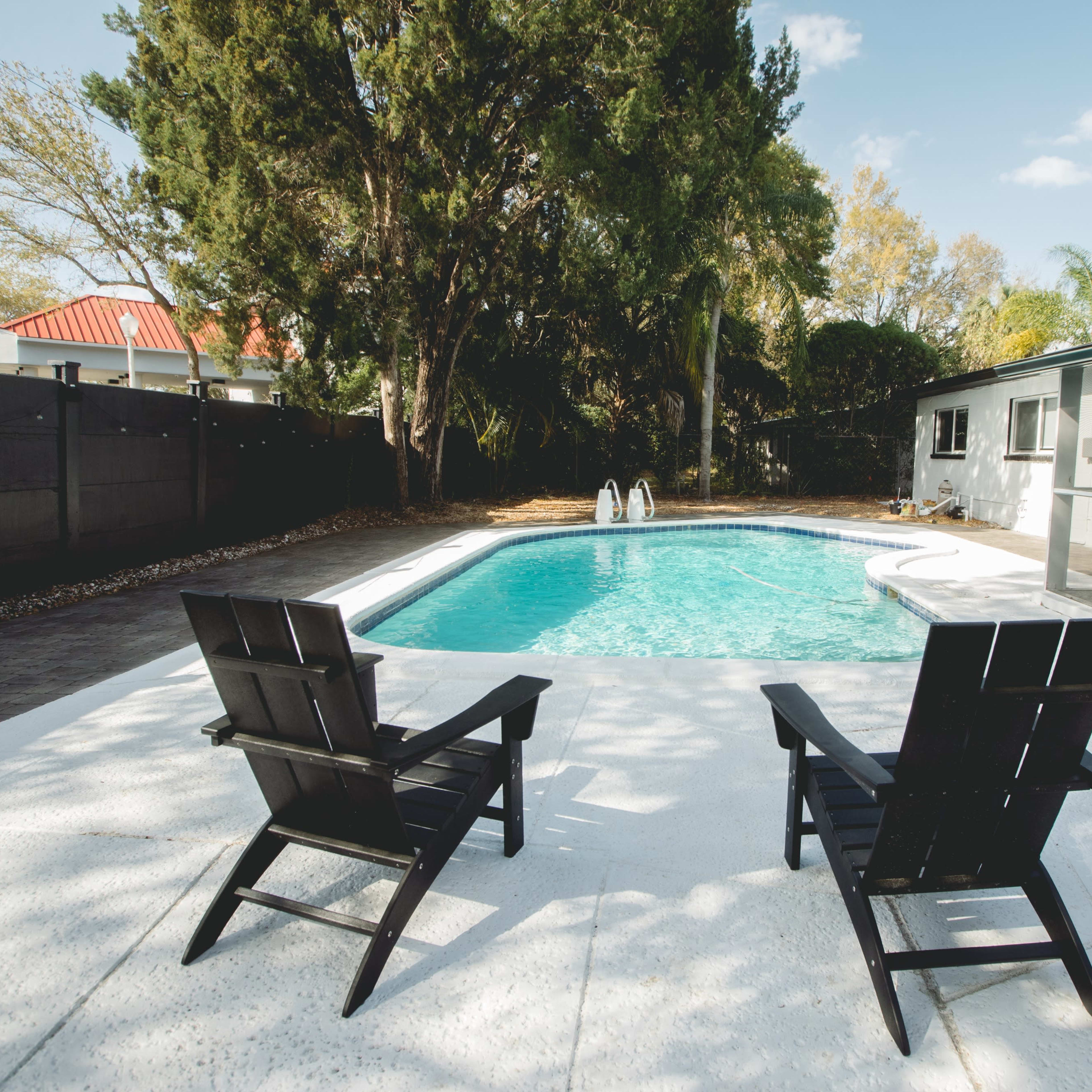 Two black outdoor chairs positioned beside a swimming pool surrounded by trees and a house.
