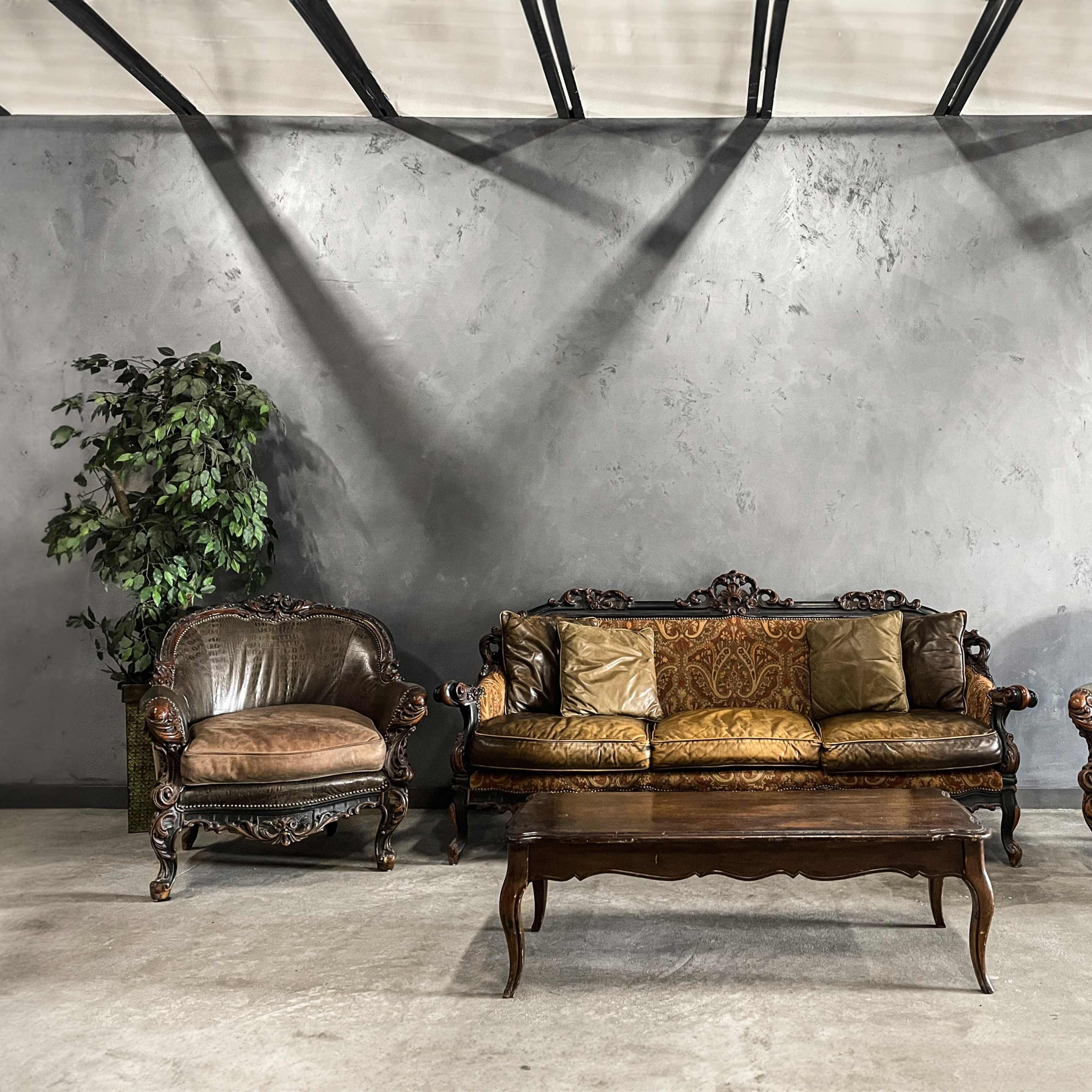 A vintage seating arrangement features a central ornate sofa flanked by two matching armchairs, set against a gray wall with a potted plant nearby.