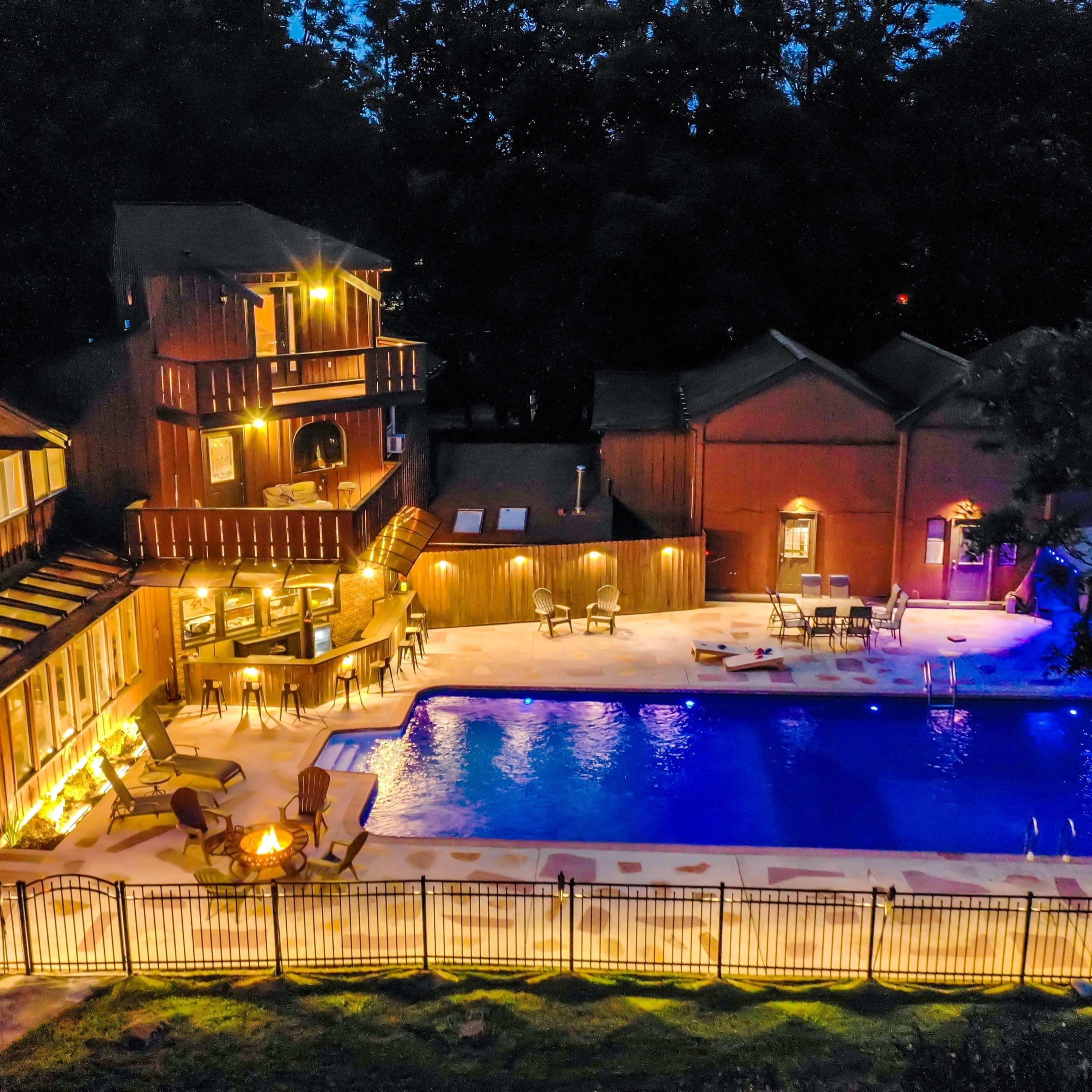A well-lit outdoor area featuring a swimming pool surrounded by chairs and tables, with multi-storied wooden buildings in the background.
