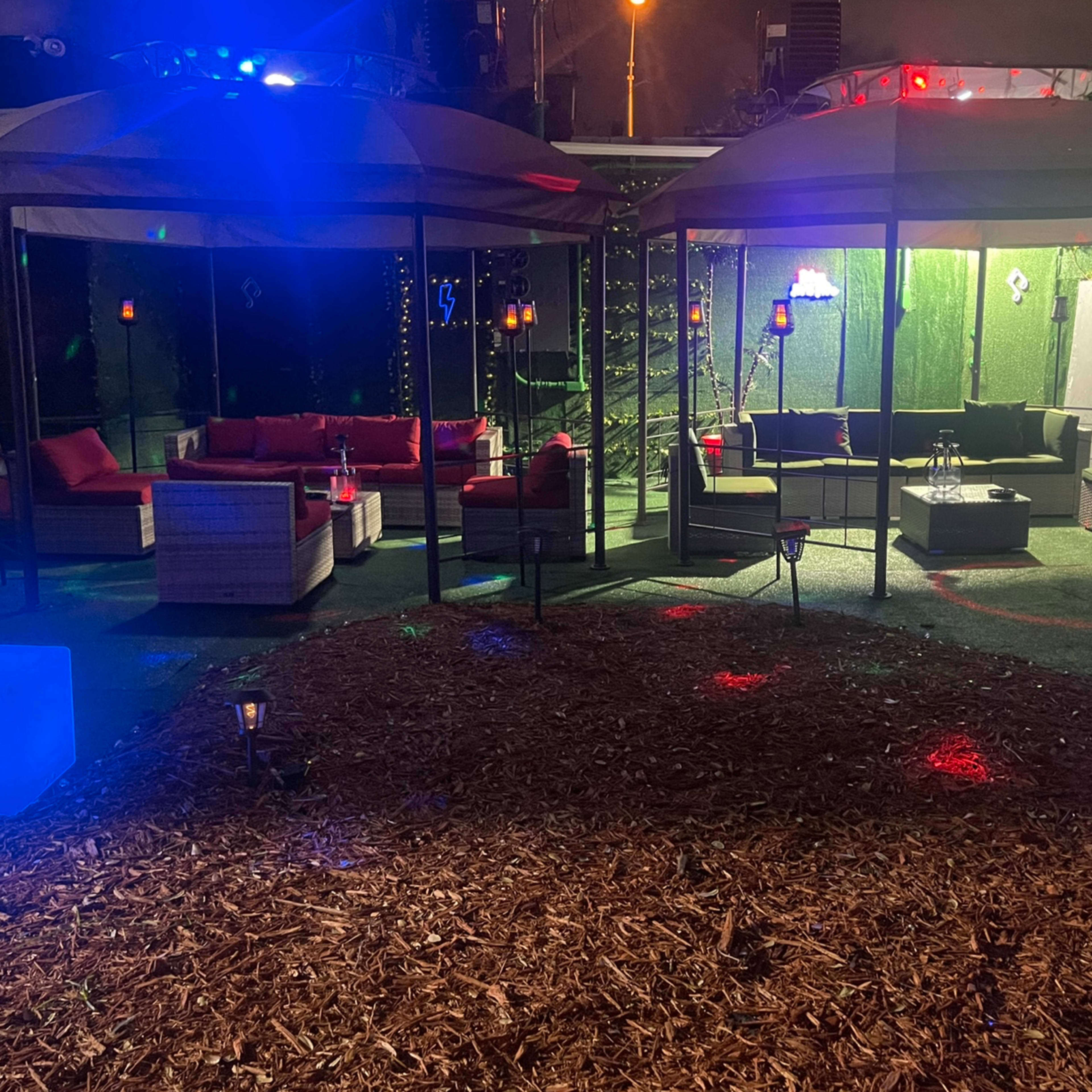A landscaped outdoor party area with two covered seating areas, decorative lights, and a woodchip ground.