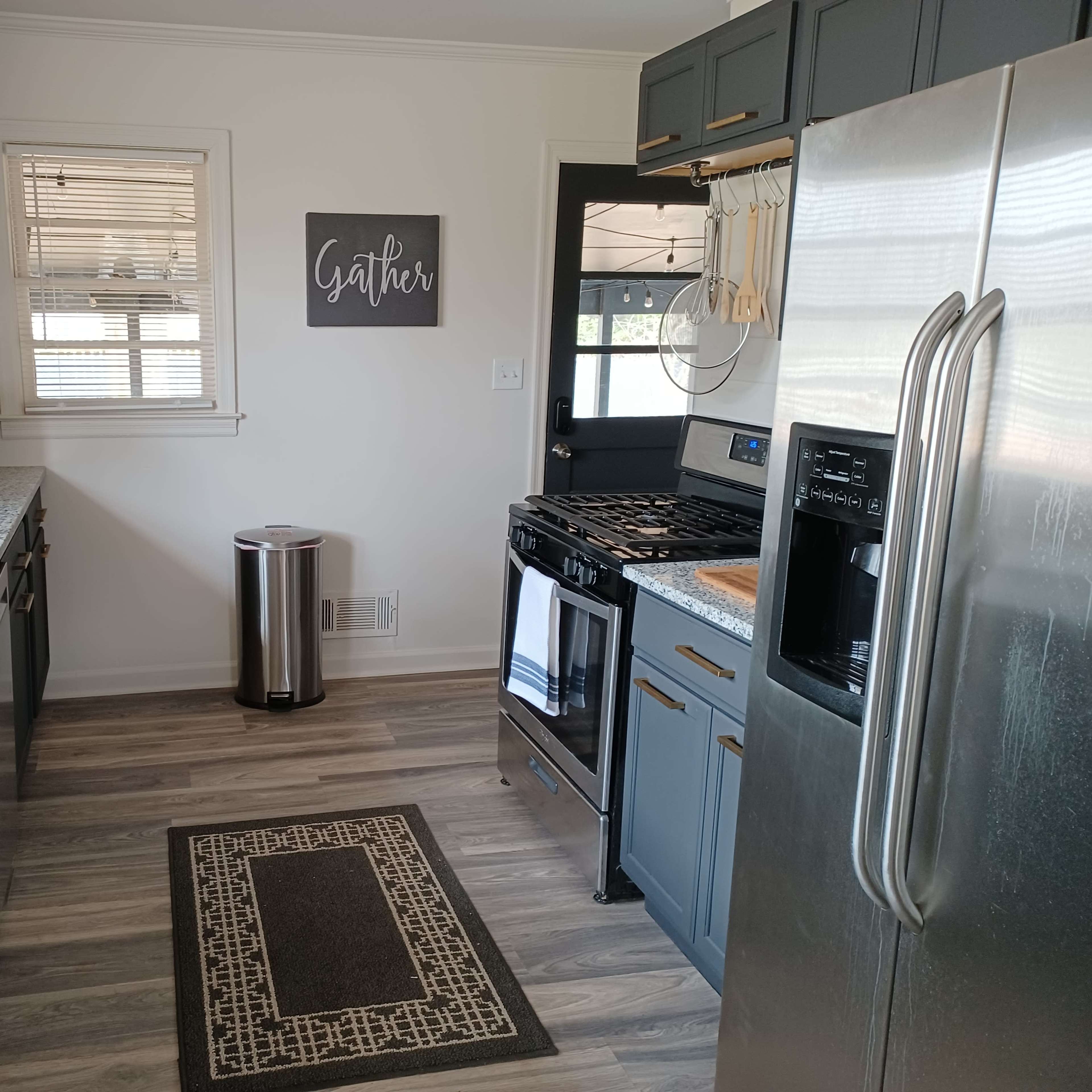 A modern kitchen features dark blue cabinets, a stainless steel refrigerator, a gas stove, and granite countertops, along with wooden shelves displaying glassware.