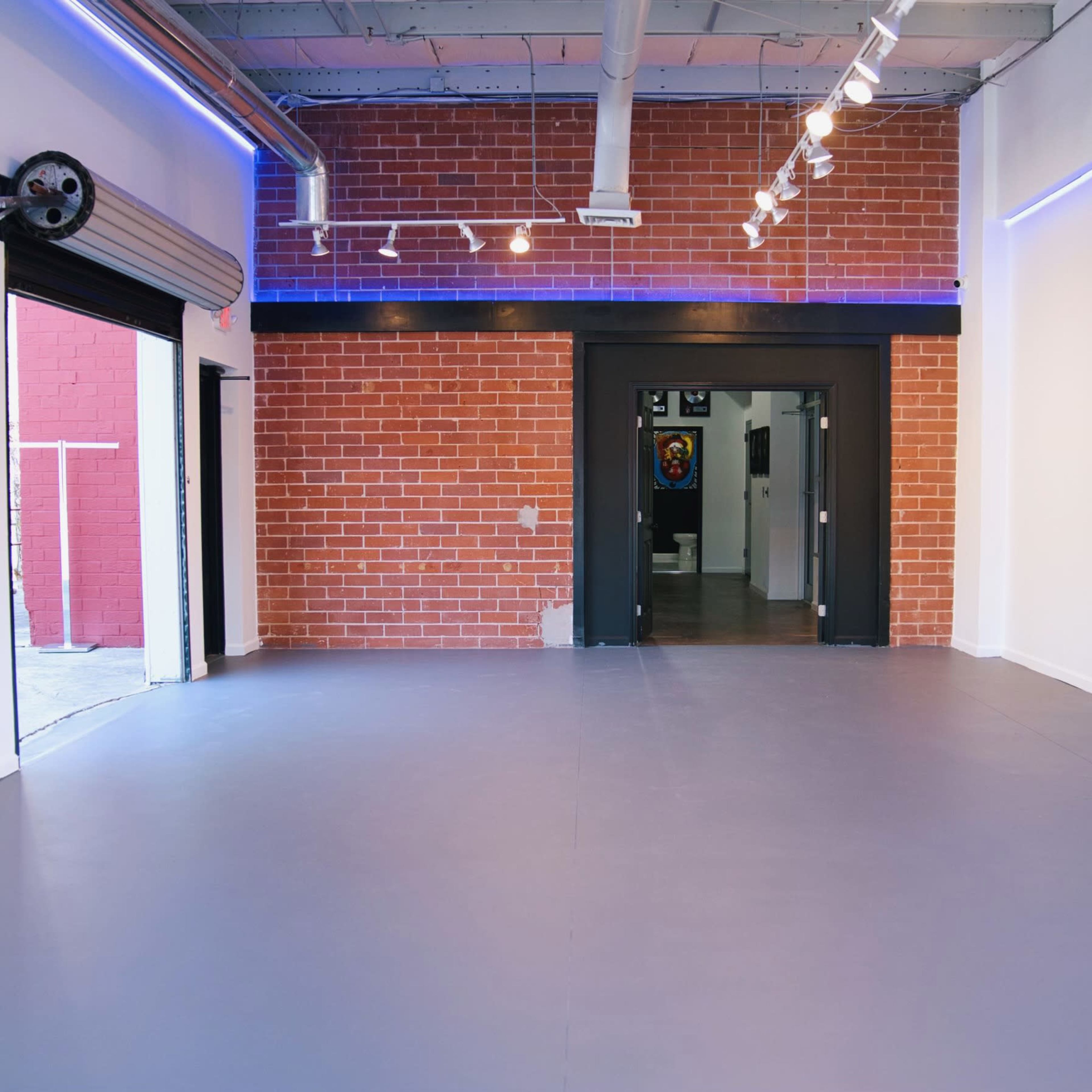 An empty interior space with a polished gray floor, exposed brick walls, and a large black door at the back leading to another room.
