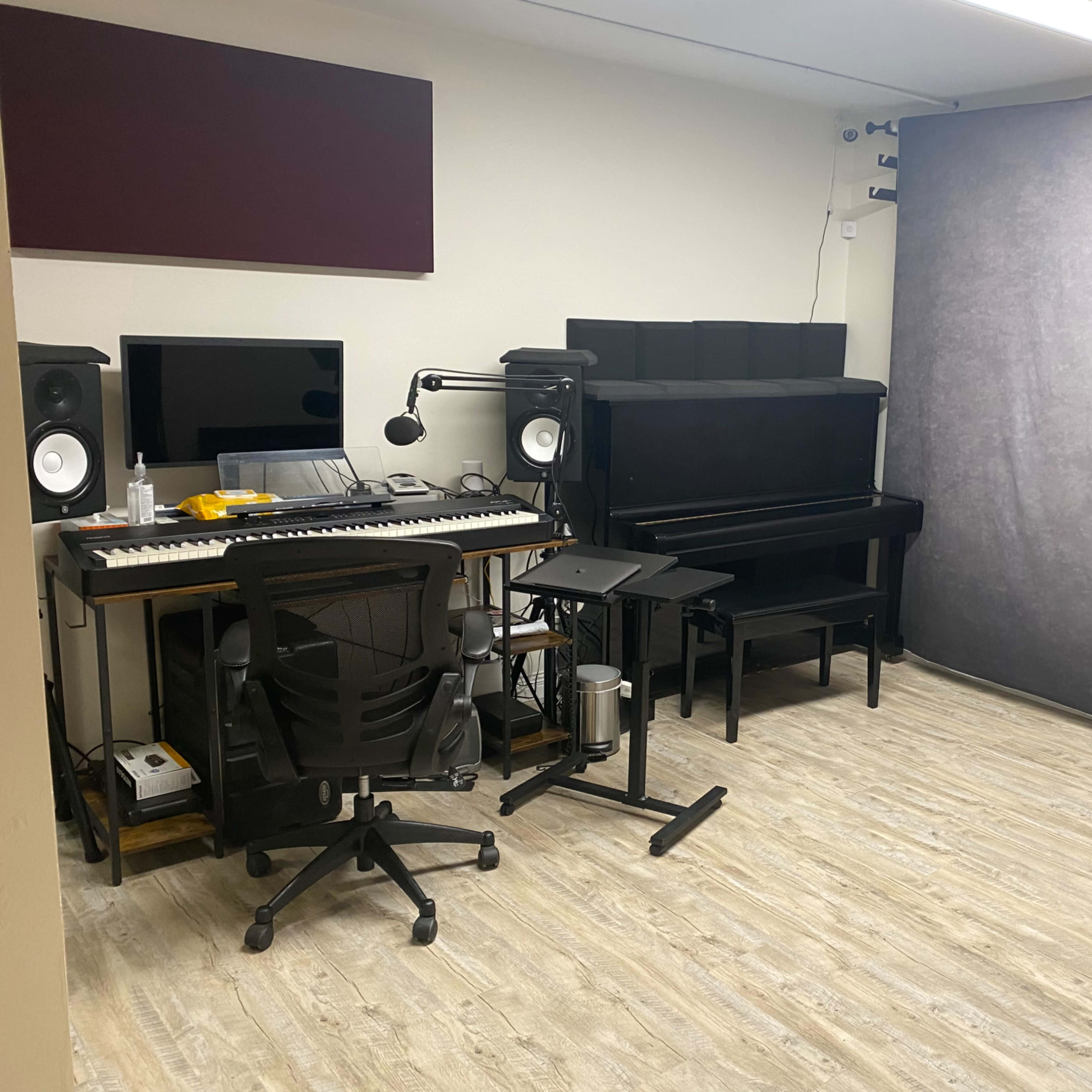A music studio featuring a computer desk with a keyboard, monitors, a microphone, a digital piano, and a backdrop.