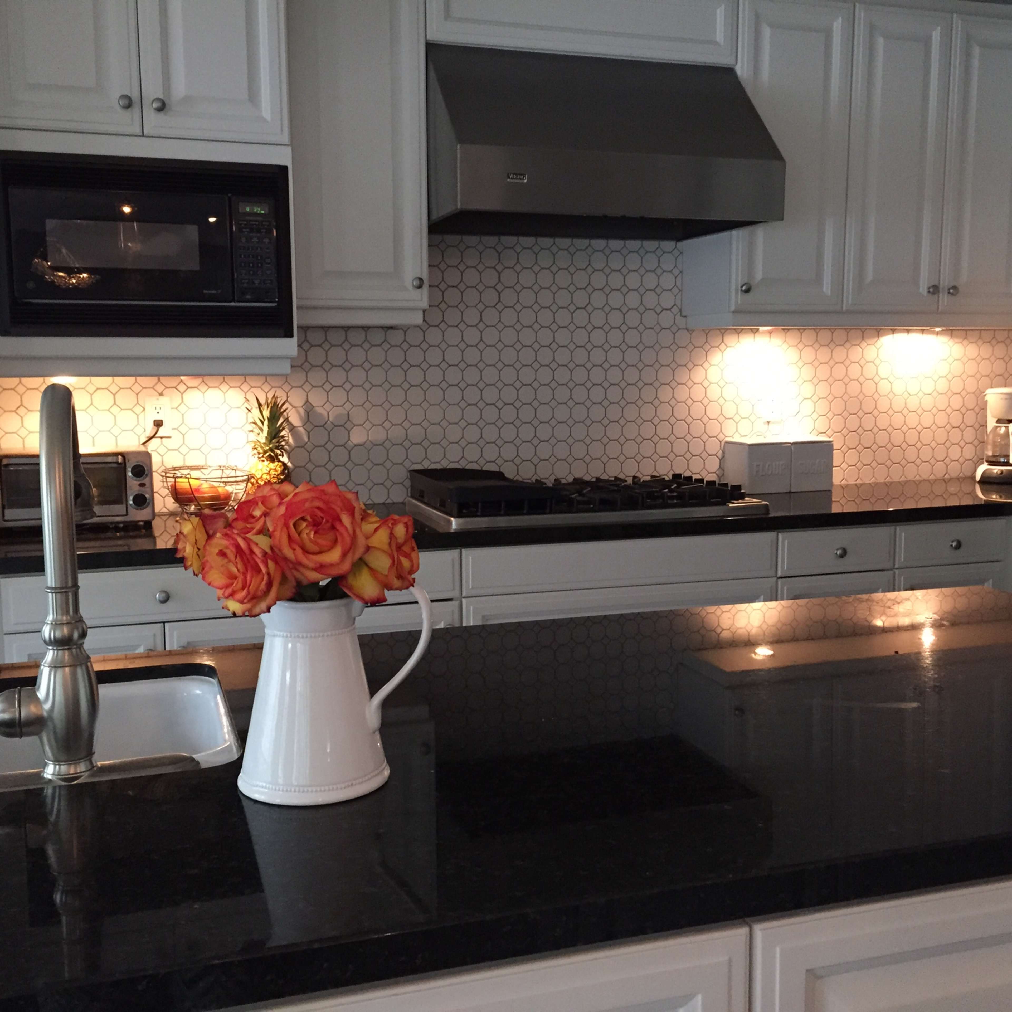 A modern kitchen features white cabinetry, a black countertop, a hexagonal backsplash, and a vase of orange roses.