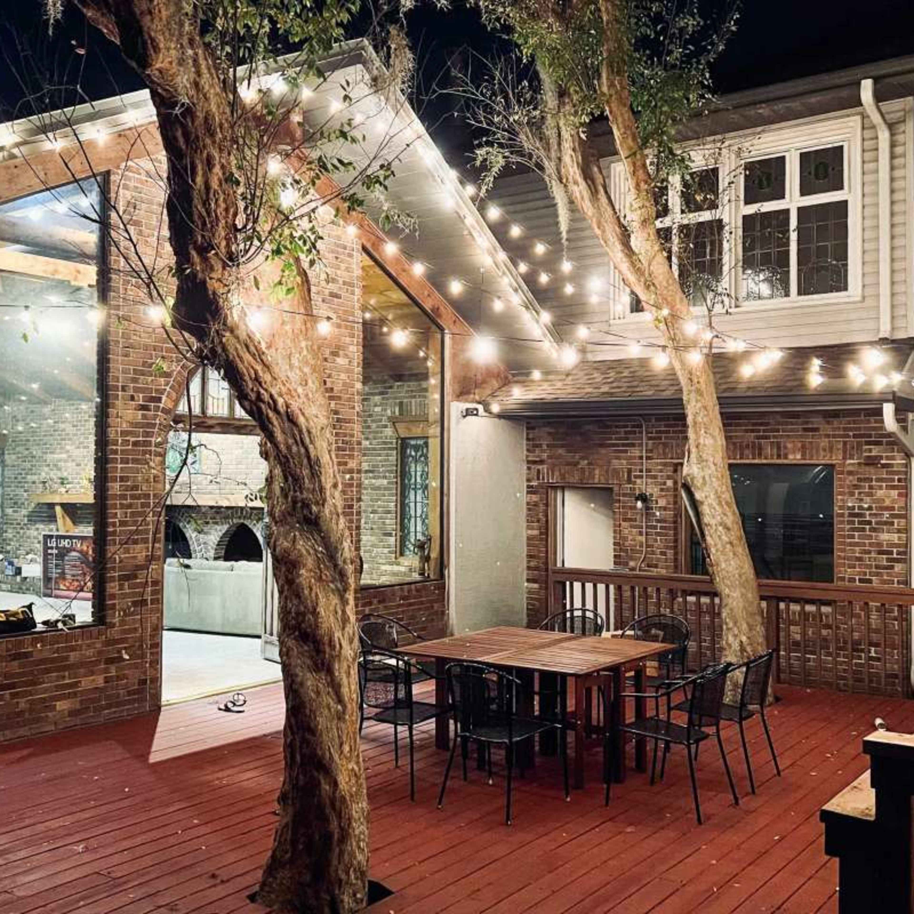 A wooden deck with a table and chairs is illuminated by string lights, adjacent to a brick house featuring large windows and visible interior spaces at night.
