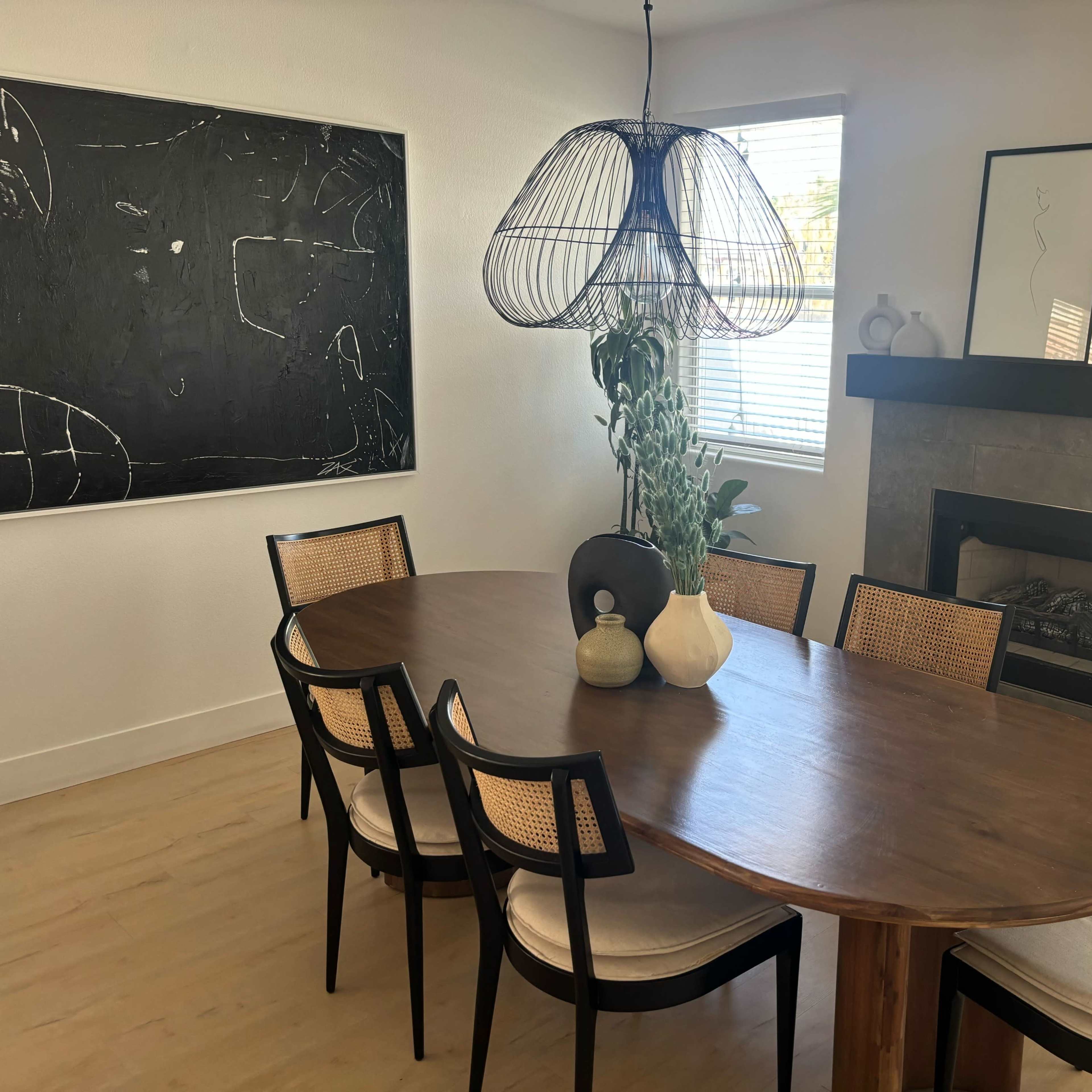 A modern dining room features a large oval wooden table surrounded by black and cane chairs, with a decorative pendant light and a stone fireplace in the background.