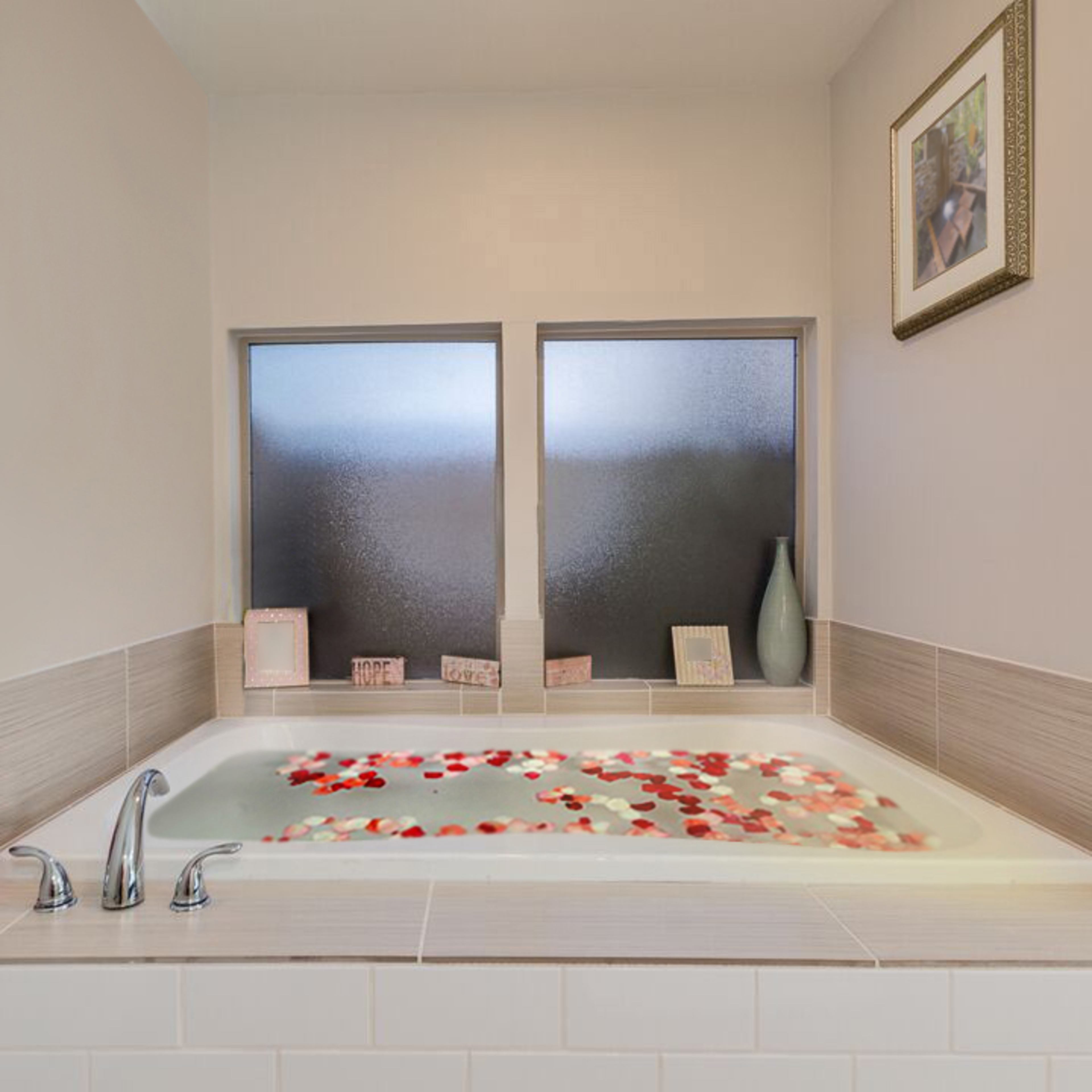 A corner bathtub filled with rose petals is positioned between two frosted windows, with framed pictures and a vase on the ledge.