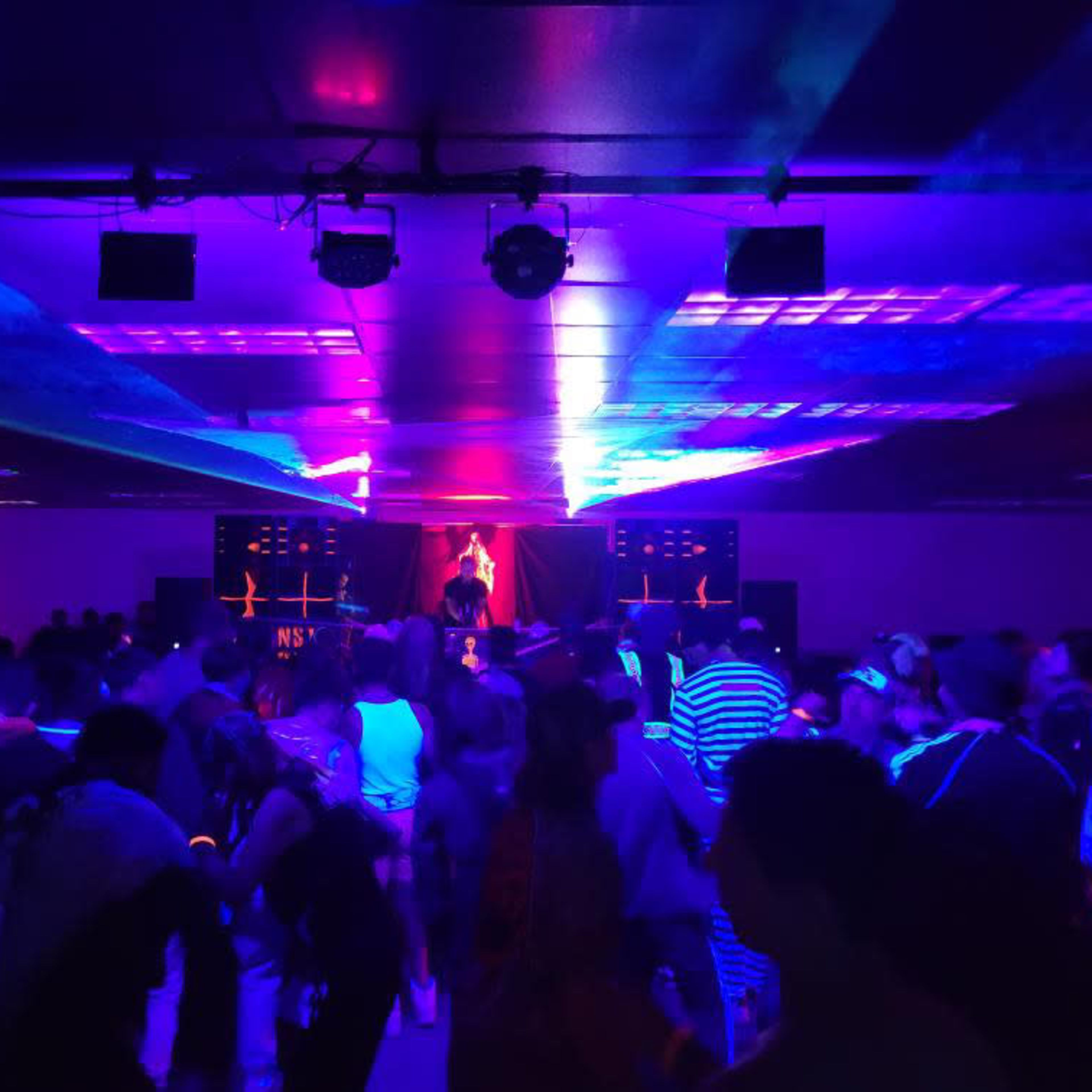 A crowded dance floor is illuminated by colorful lights, with a performer on stage in the background.