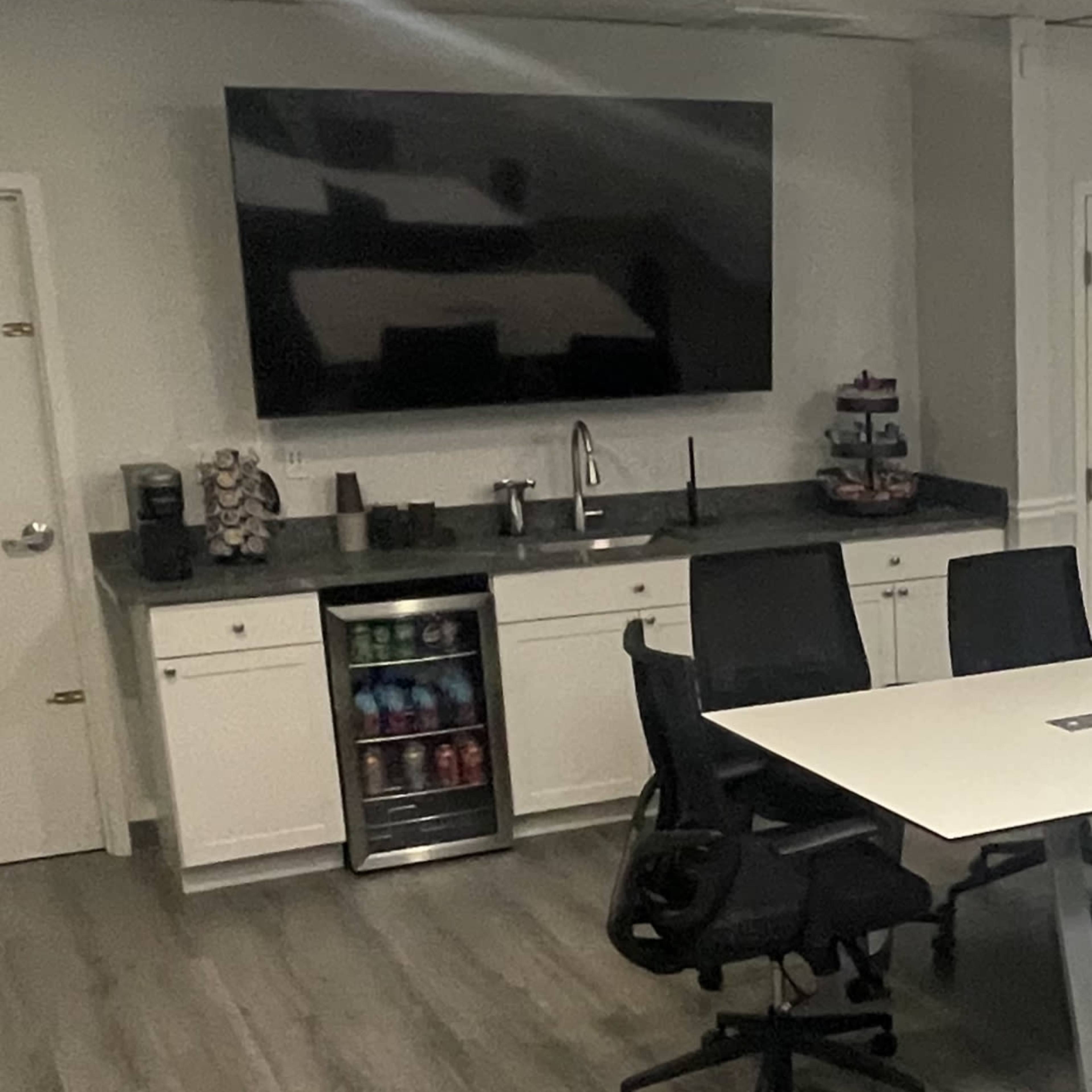 A modern office break area featuring a large wall-mounted television, a kitchen counter with a sink, a mini-fridge displaying beverages, and a seating area with black office chairs around a table.