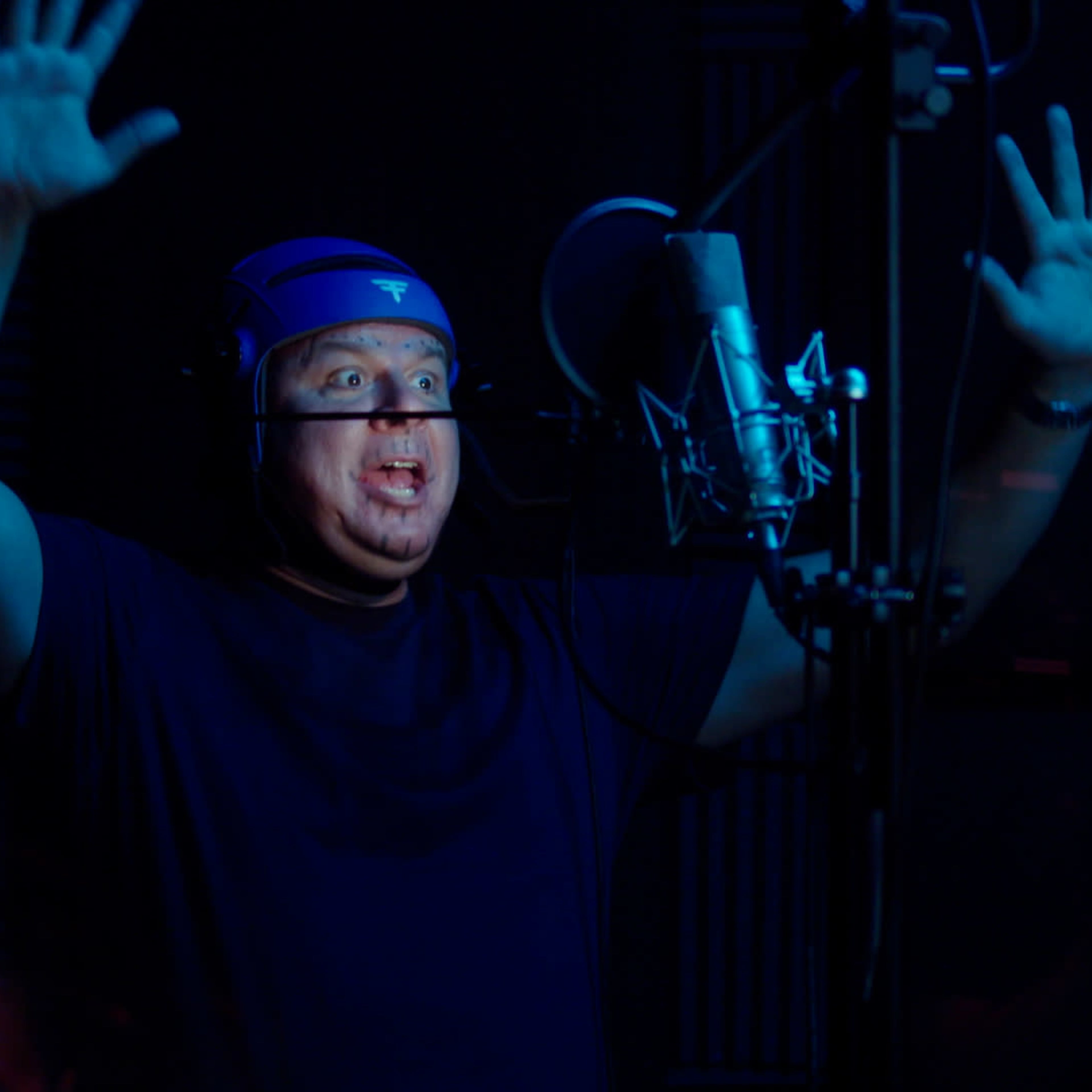 A man with a blue headband is performing in a recording studio while standing in front of a microphone with his hands raised.