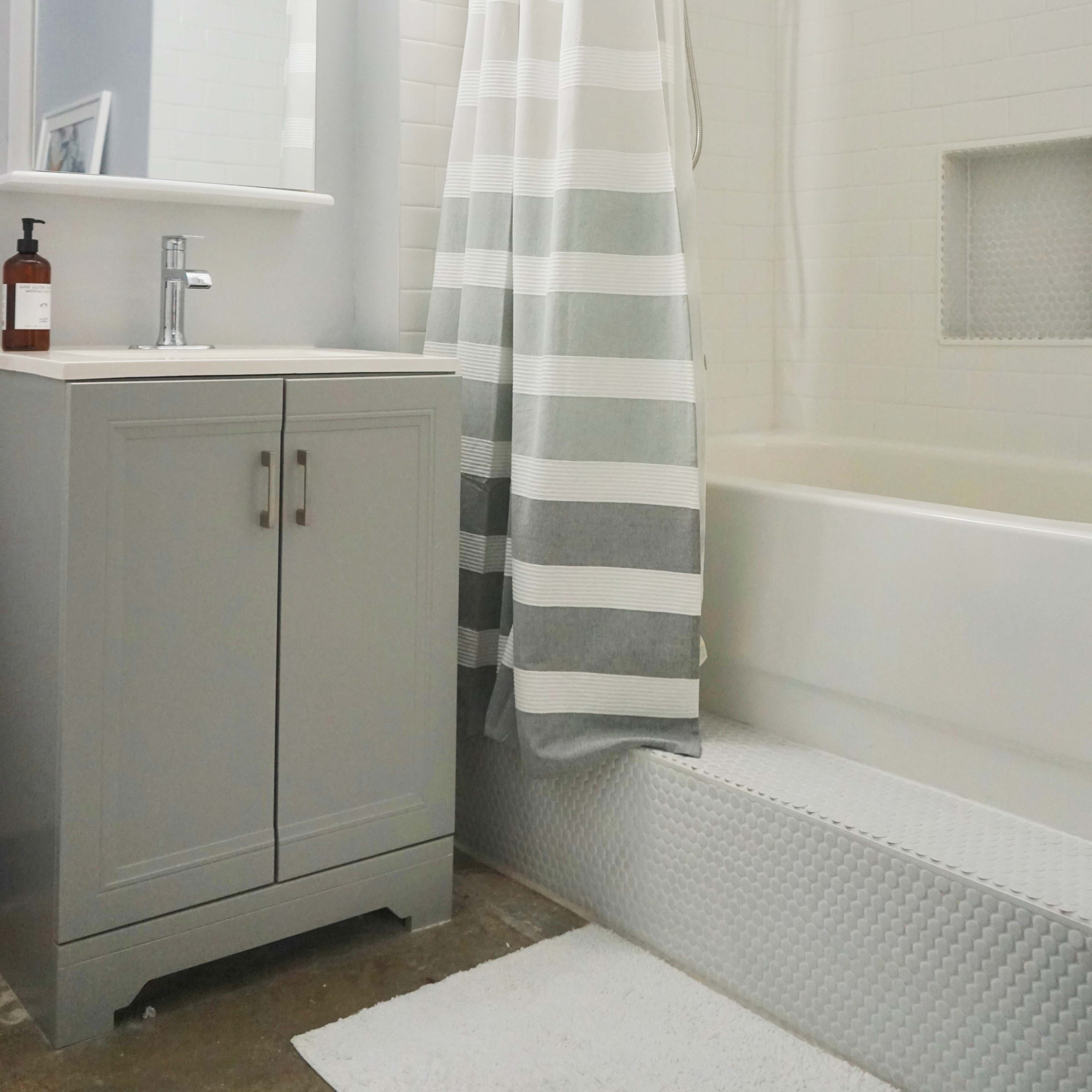 A modern bathroom features a shower with a striped curtain, a white bathtub, and a gray vanity with a mirror above it.