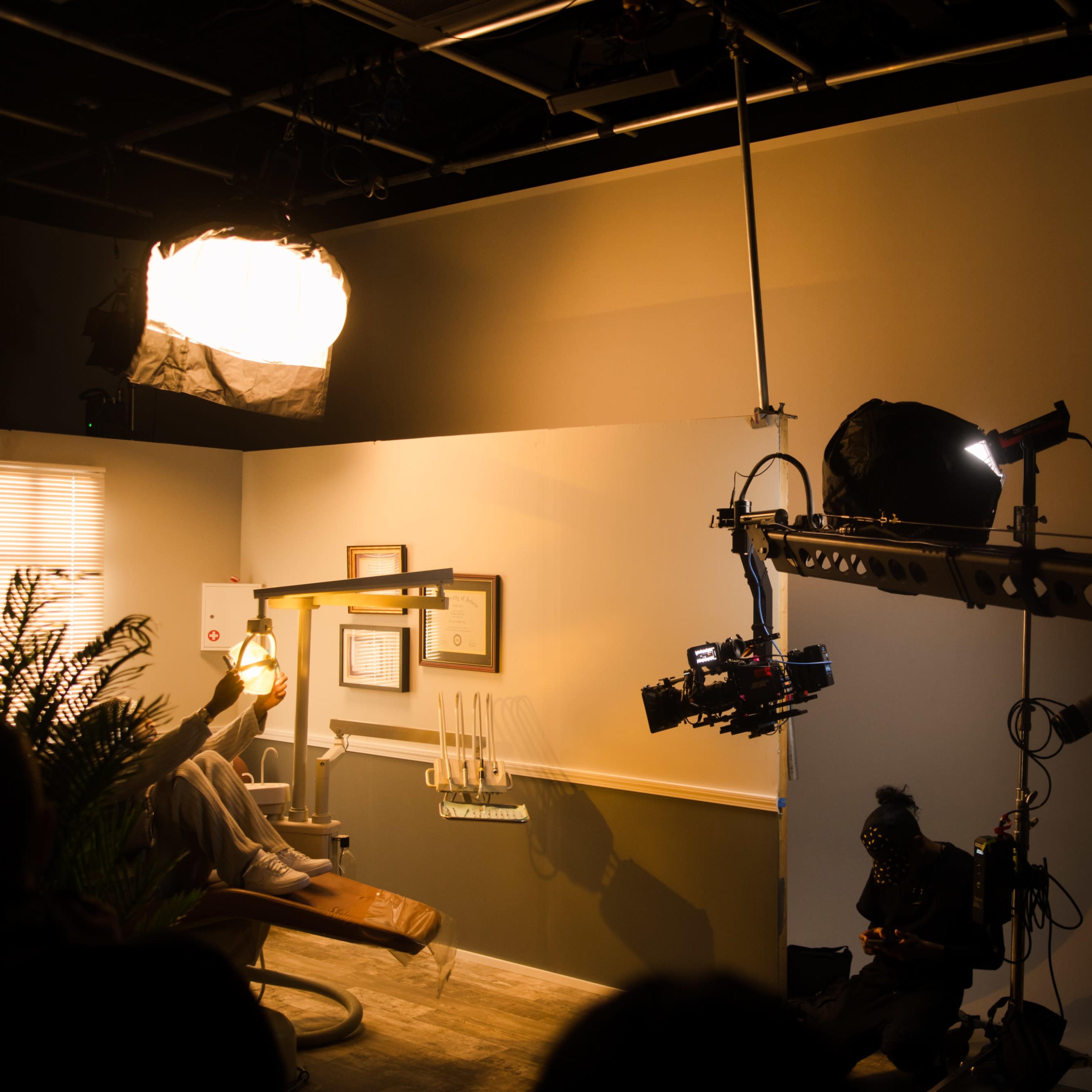 A film crew sets up equipment in a dental office for a shoot, with a camera on a crane and lights positioned overhead.