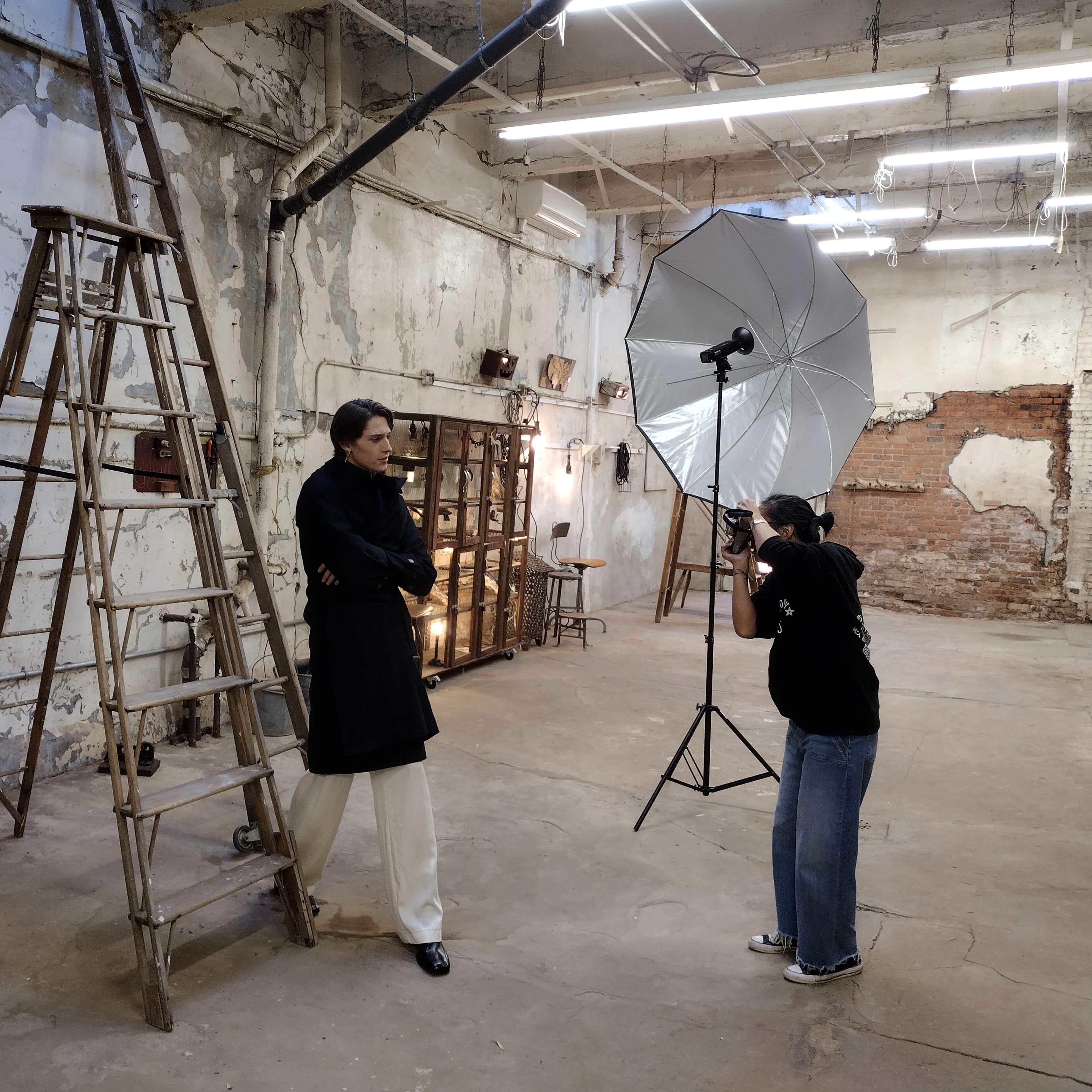 A photographer is capturing images of a model standing next to a ladder in an industrial-style studio with exposed brick walls.