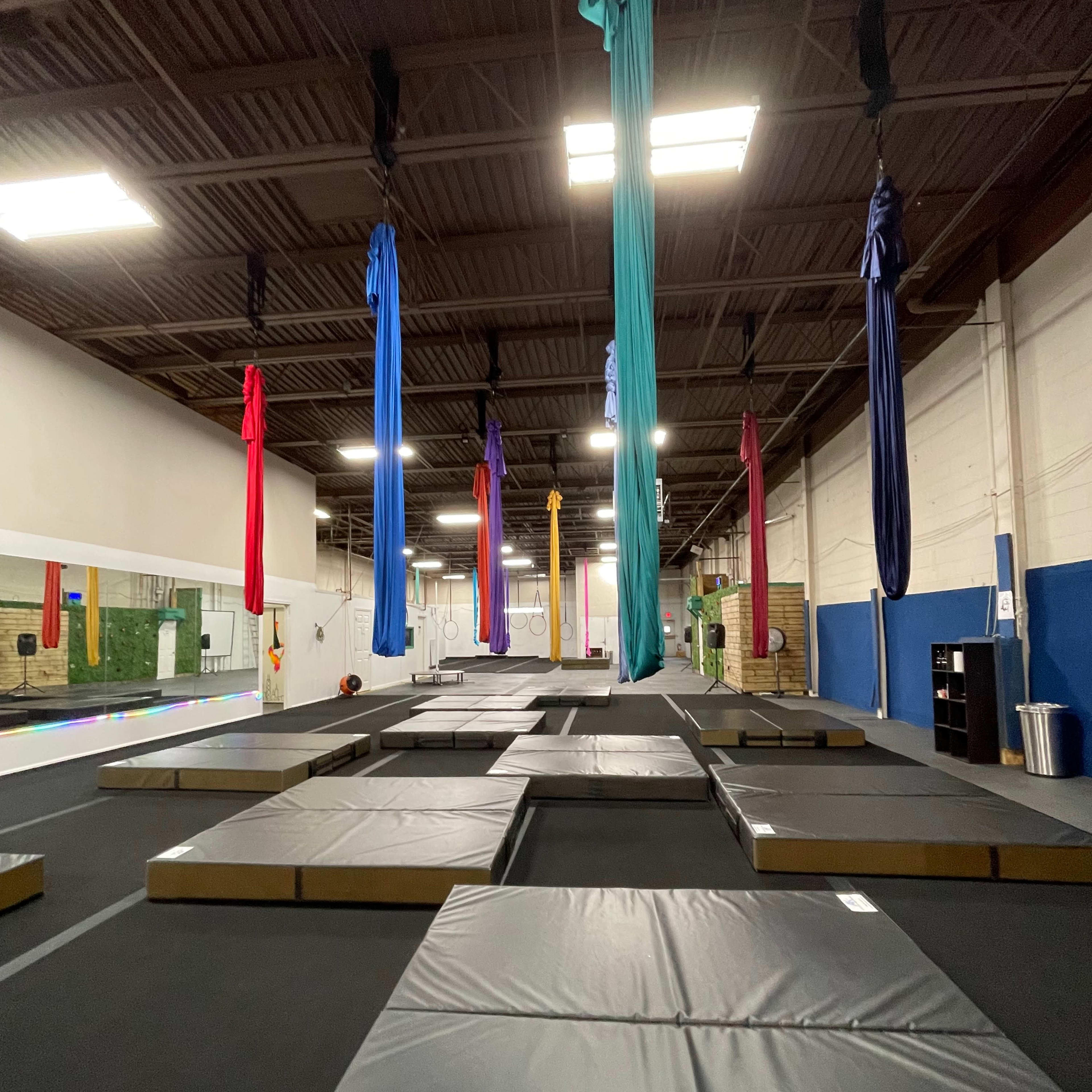 An indoor gym space with several large, padded mats and colorful aerial silks hanging from the ceiling.
