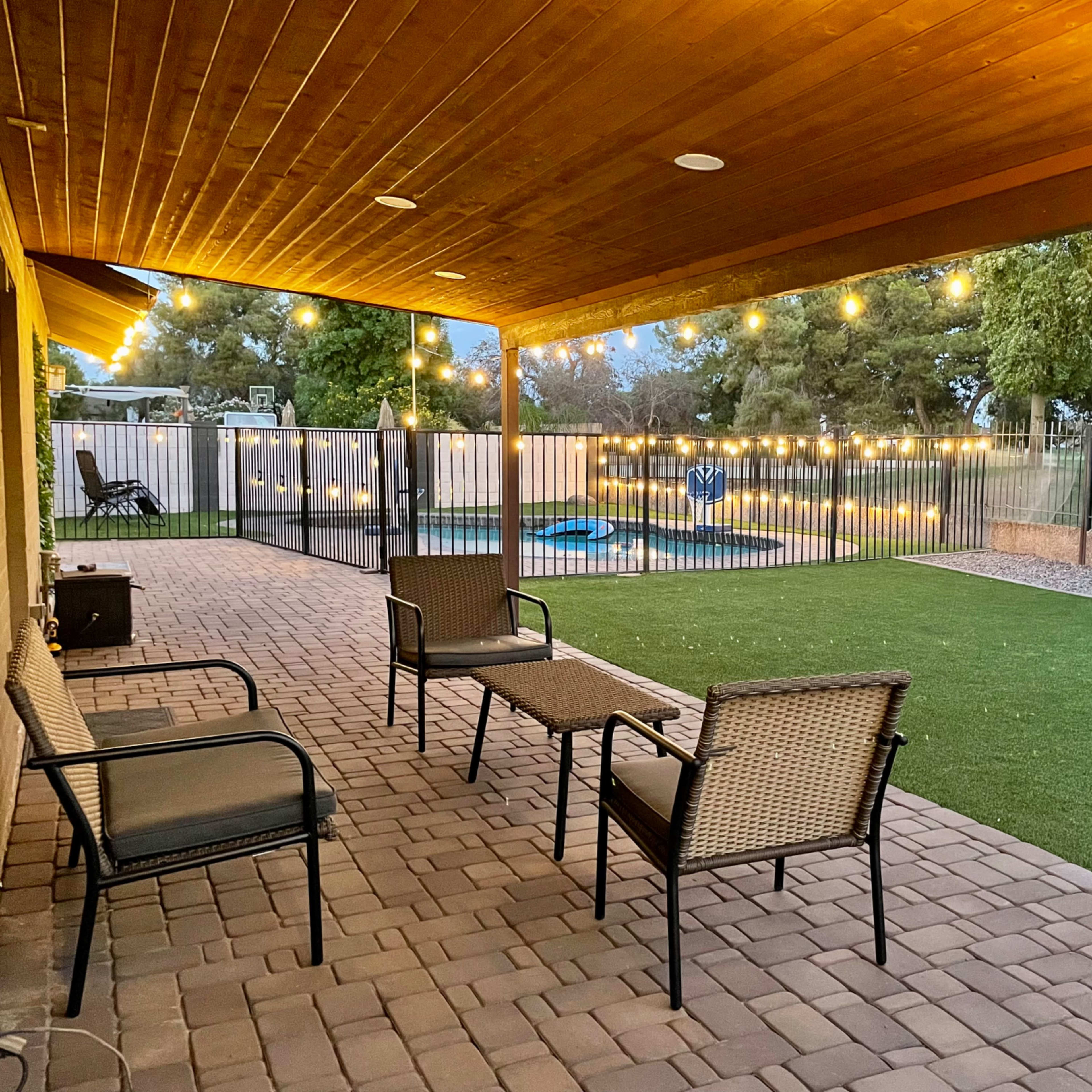 A covered patio features several chairs and a small table, illuminated by string lights, overlooking a fenced yard with a pool and greenery.