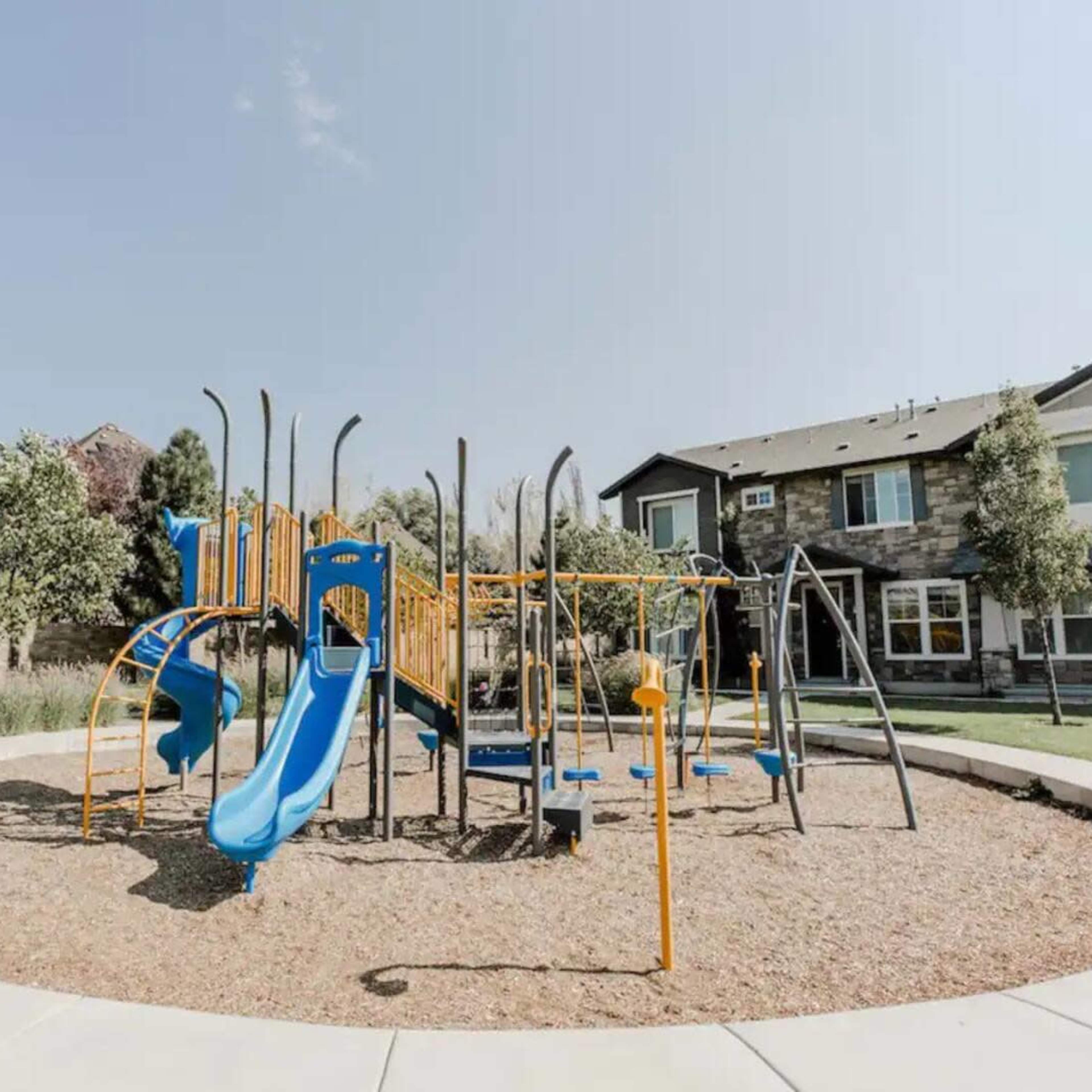 A playground with blue and orange equipment, including slides and climbing structures, situated on a gravel surface beside a modern house.