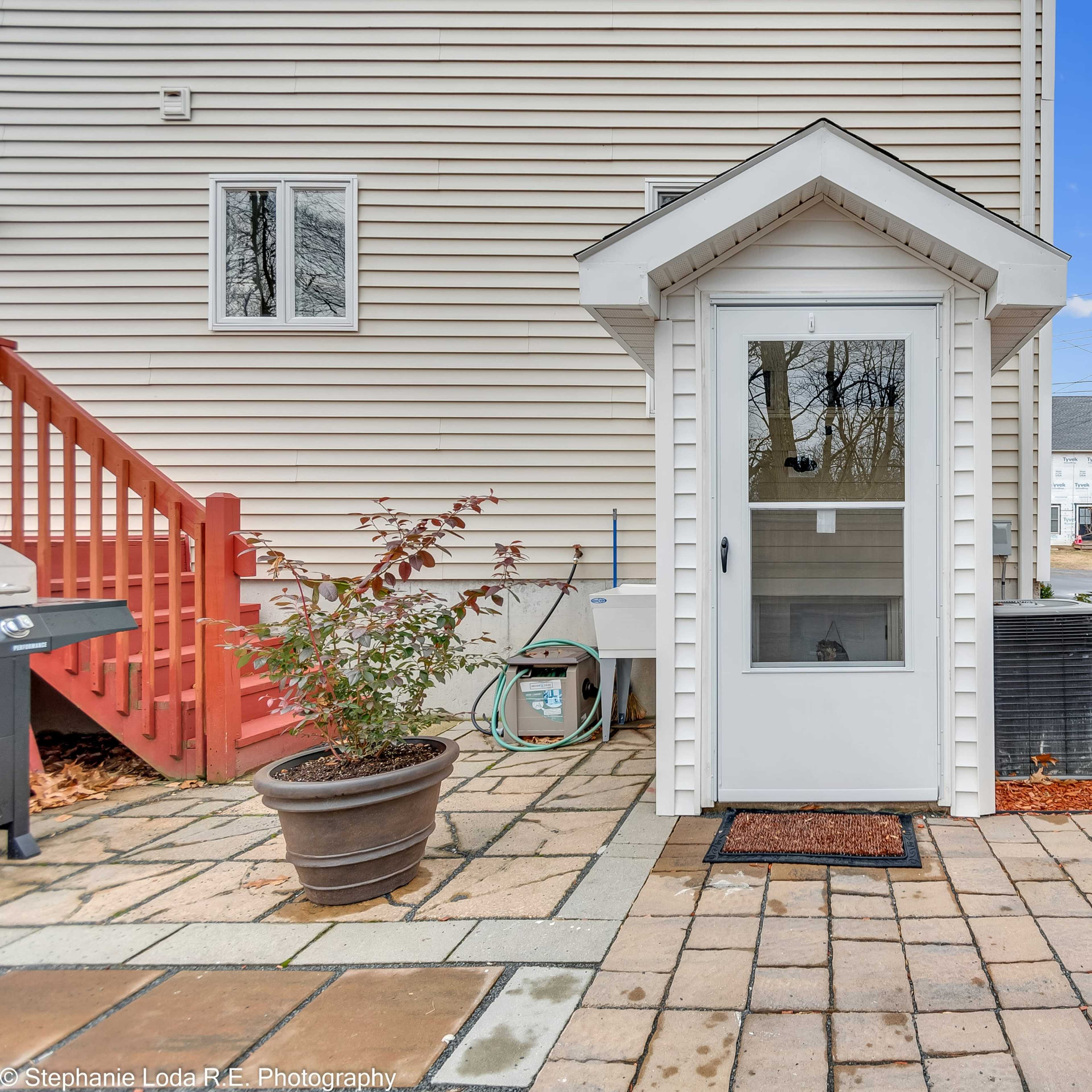 A small, white wooden shed with a peaked roof stands on a paved patio beside a fenced yard.