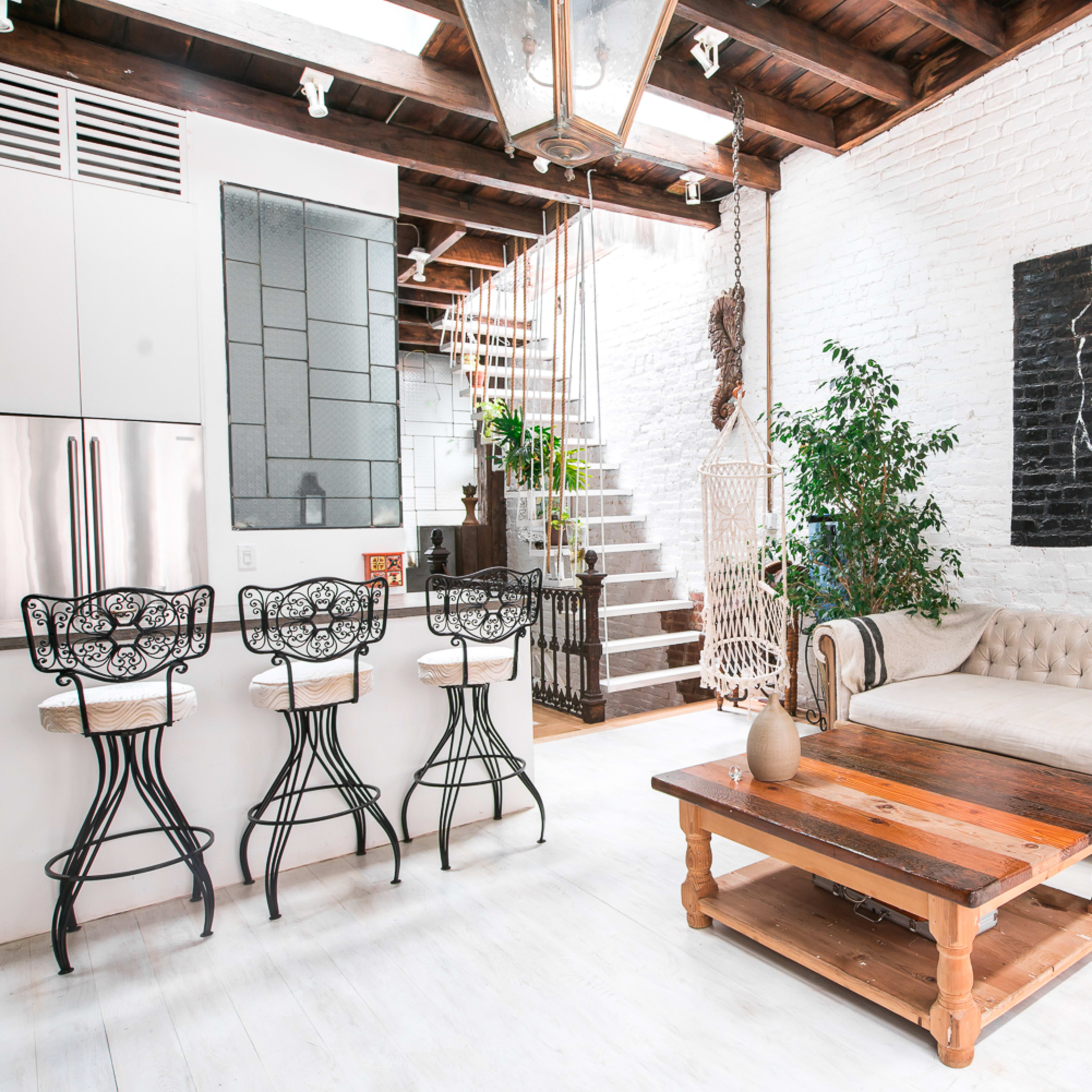 A modern living space features a white brick wall, a beige sofa, a wooden coffee table, and bar stools at a kitchen counter, with a staircase in the background leading to an upper level.