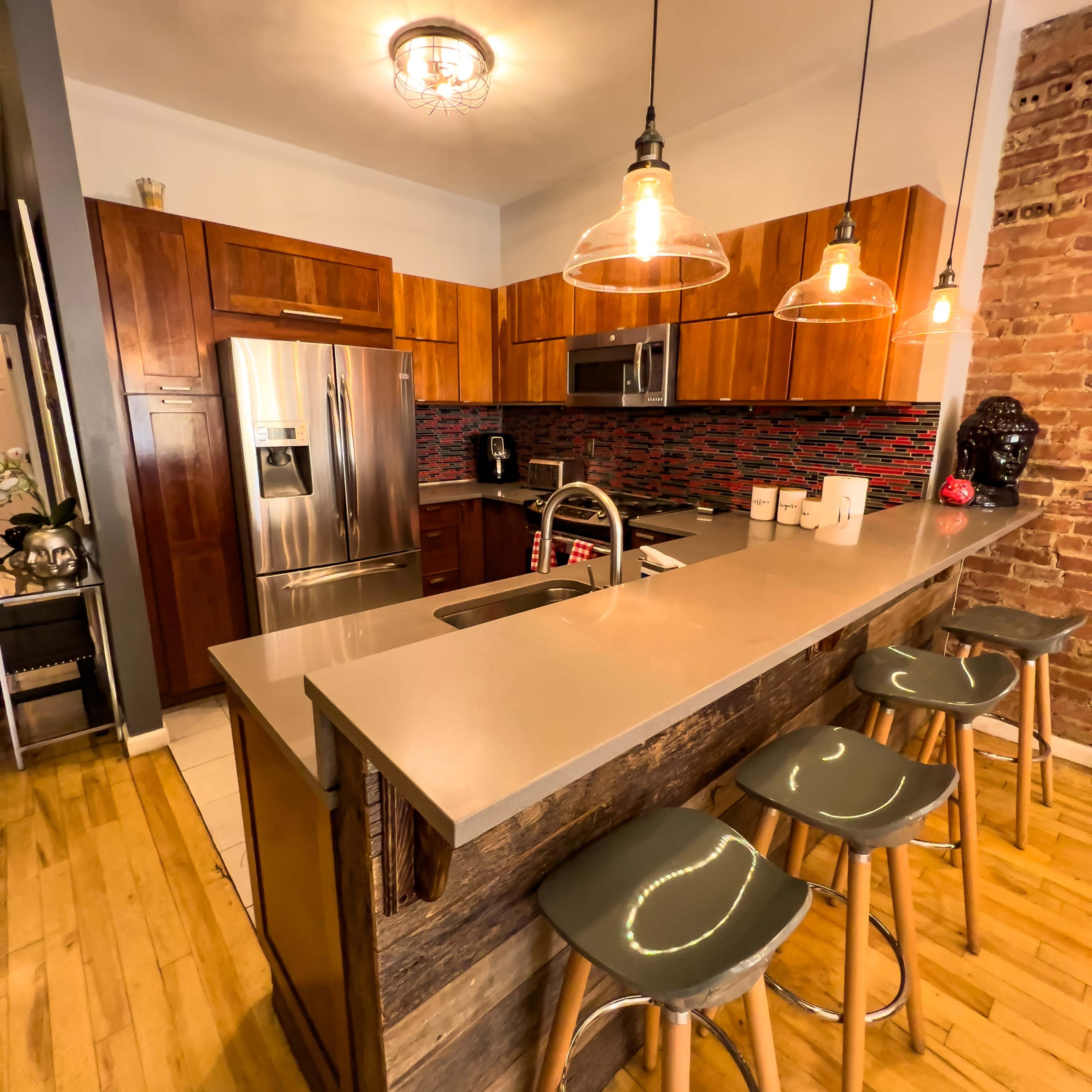 A modern kitchen with wooden cabinets, a gray countertop, an island with three stools, and exposed brick walls.