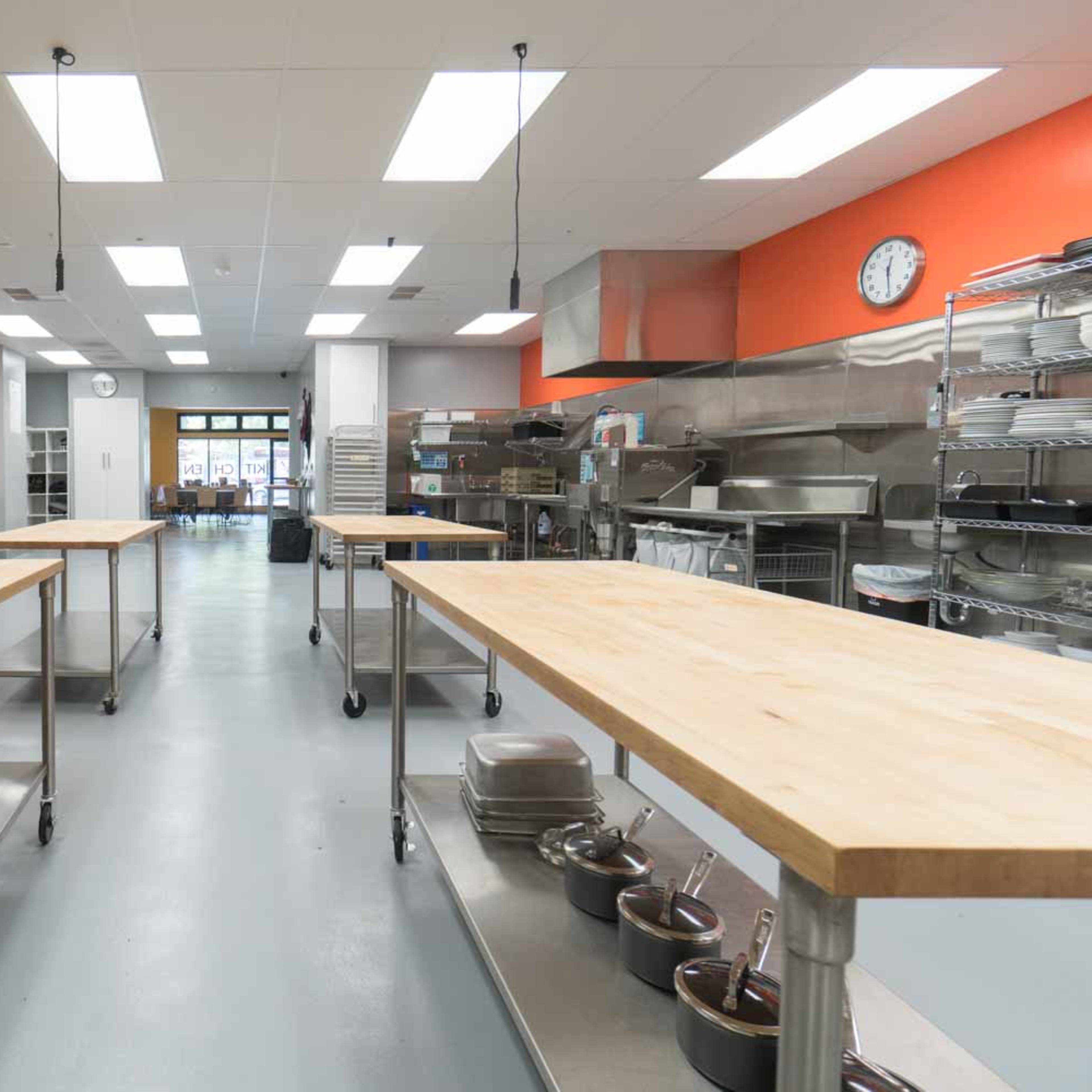 A bright commercial kitchen with stainless steel appliances and wooden worktables arranged in a spacious layout.