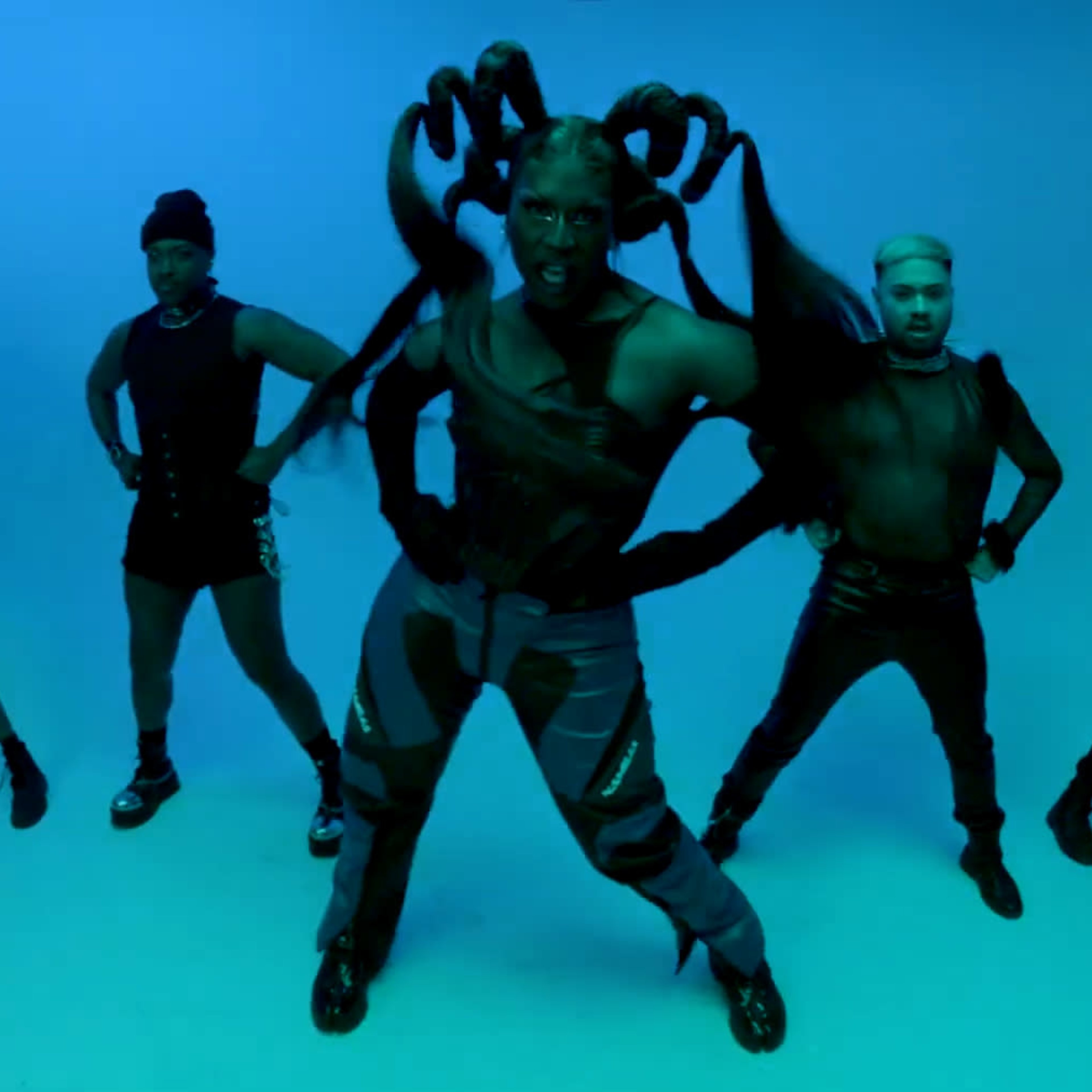 Five performers pose against a blue background, showcasing a variety of stylish outfits and creative hairstyles, with one individual exhibiting elaborate, tentacle-like hair extensions.