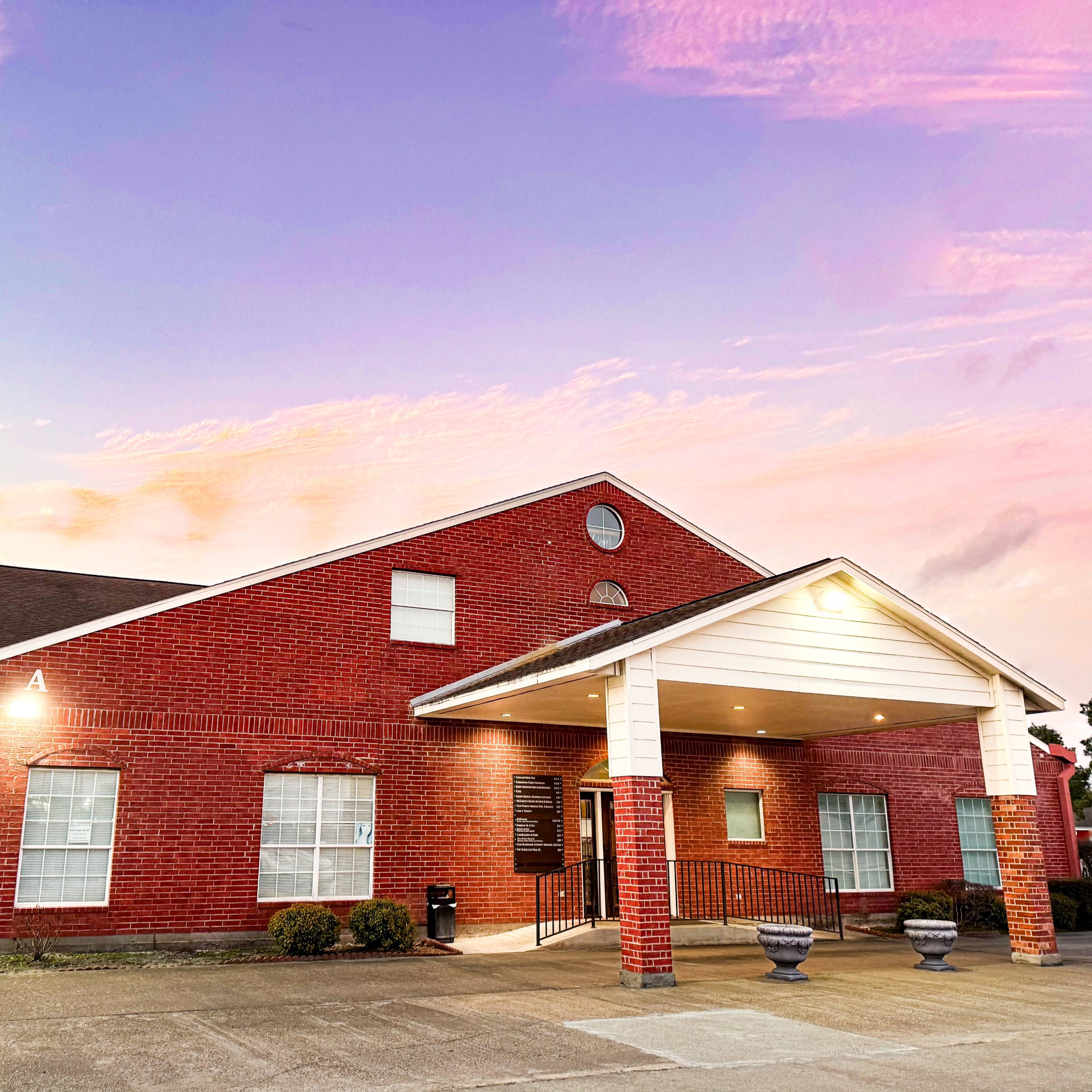 A red brick building with a white entrance and landscaping, set against a colorful sunset sky.