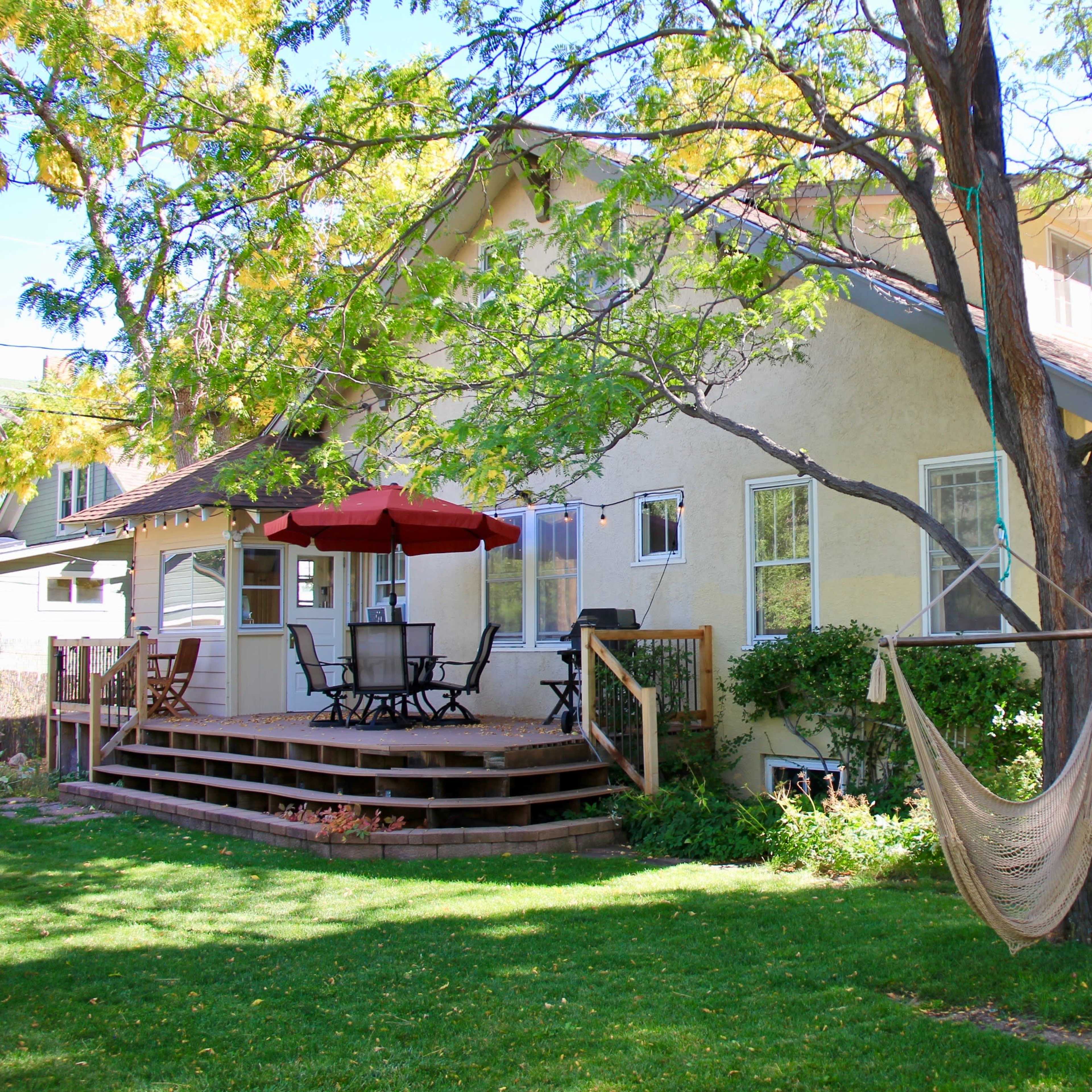 A light-colored house with a wooden deck features a hammock hanging from a tree and seating under an umbrella in the backyard.