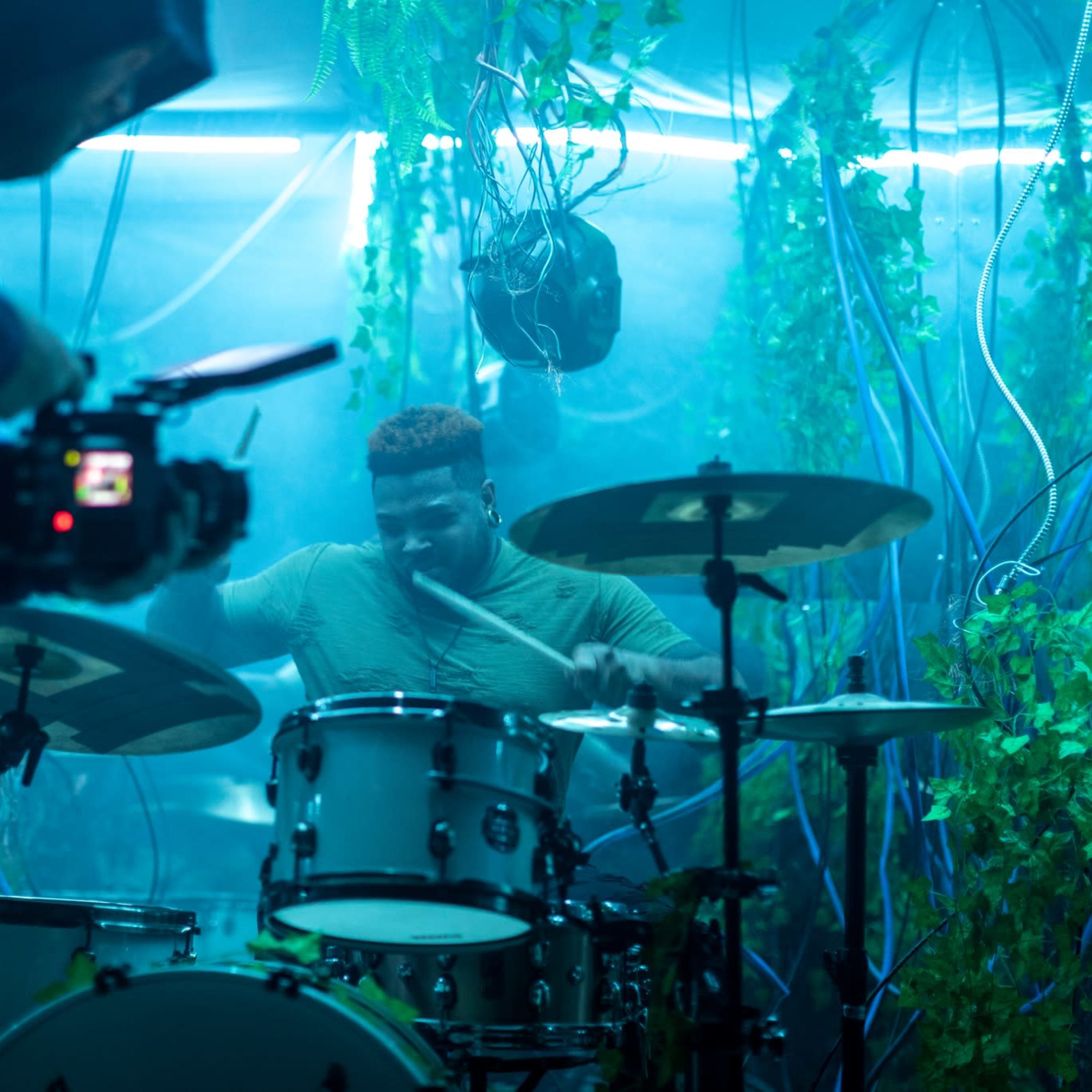 A drummer performs on a white drum set under blue lights in a setting decorated with green vines and wires, while a cameraman films him.