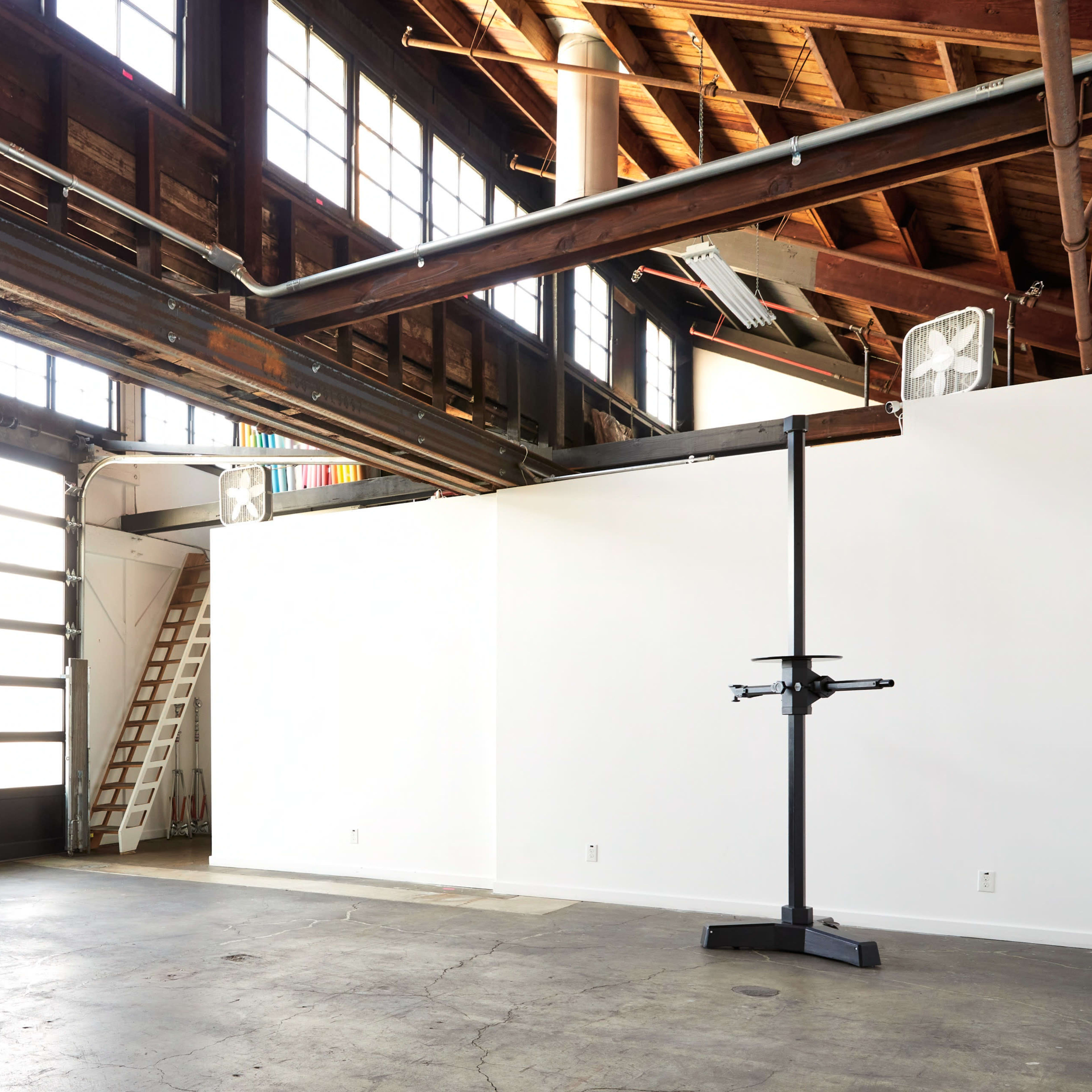A spacious industrial-style room with large windows, a high ceiling, and a loading dock door, featuring a blank white wall and a mounted display stand.