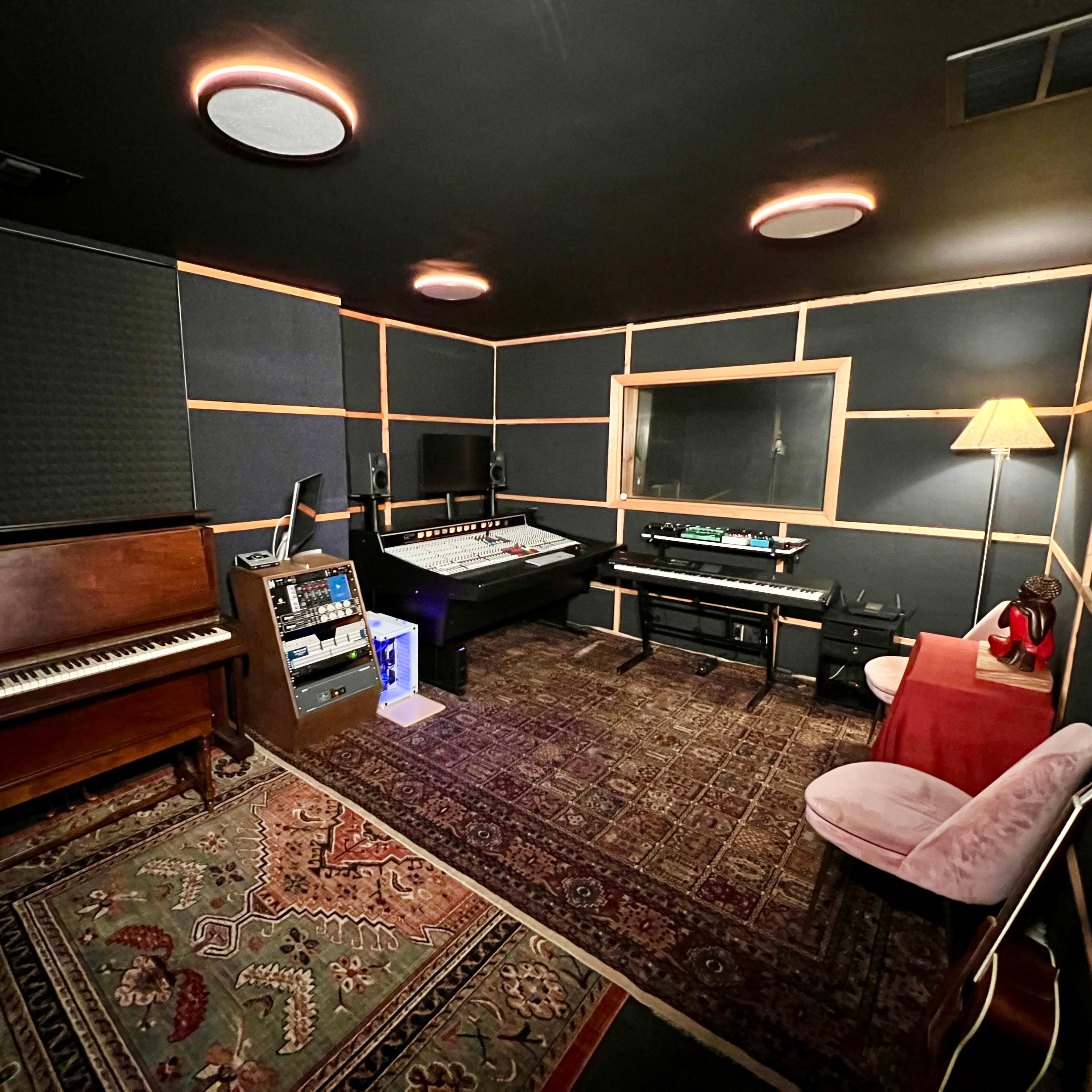 A music studio with a piano, electronic equipment, a chair, and a lamp arranged around a carpeted floor.