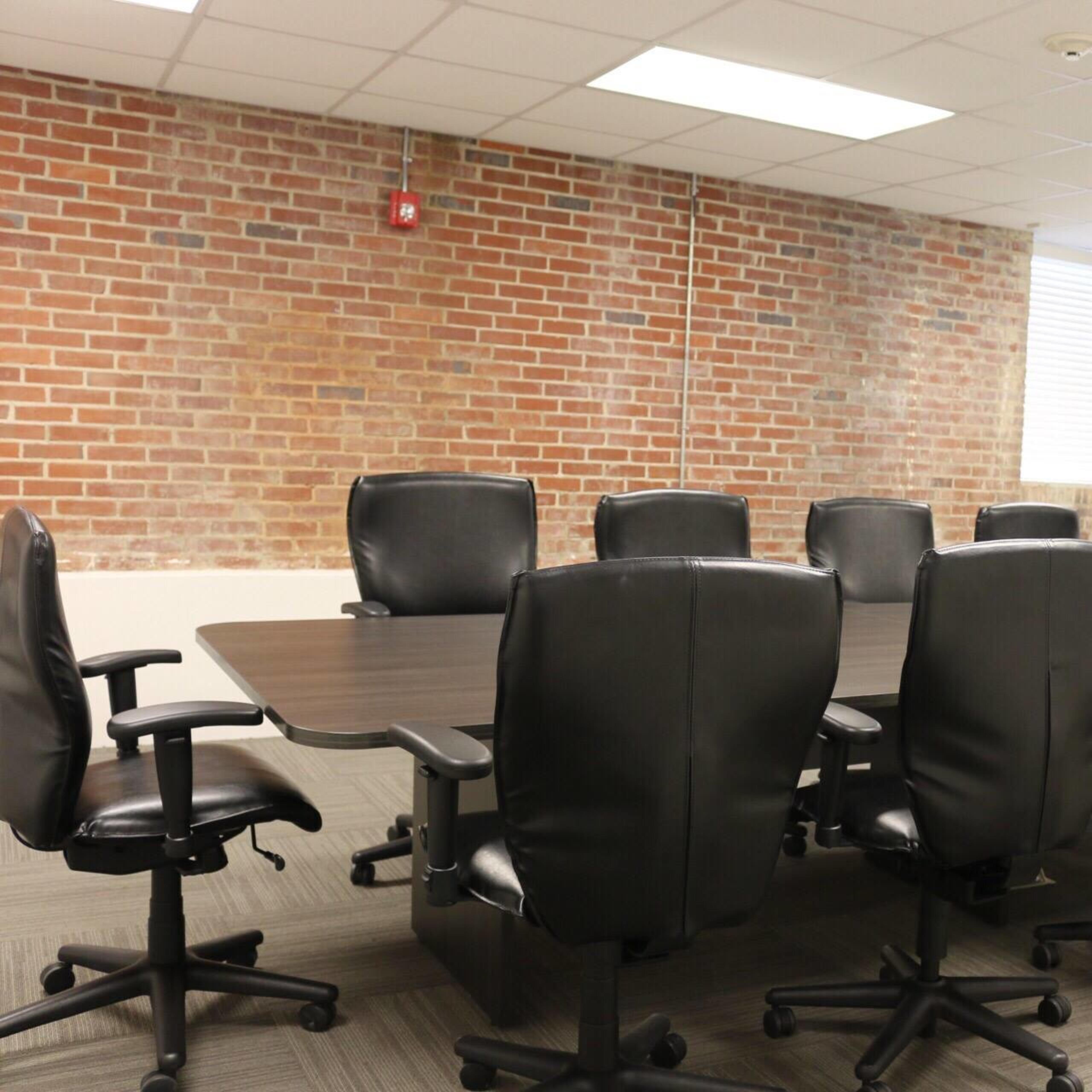 A conference room with a long, dark wooden table surrounded by ergonomic black chairs, set against a brick wall.