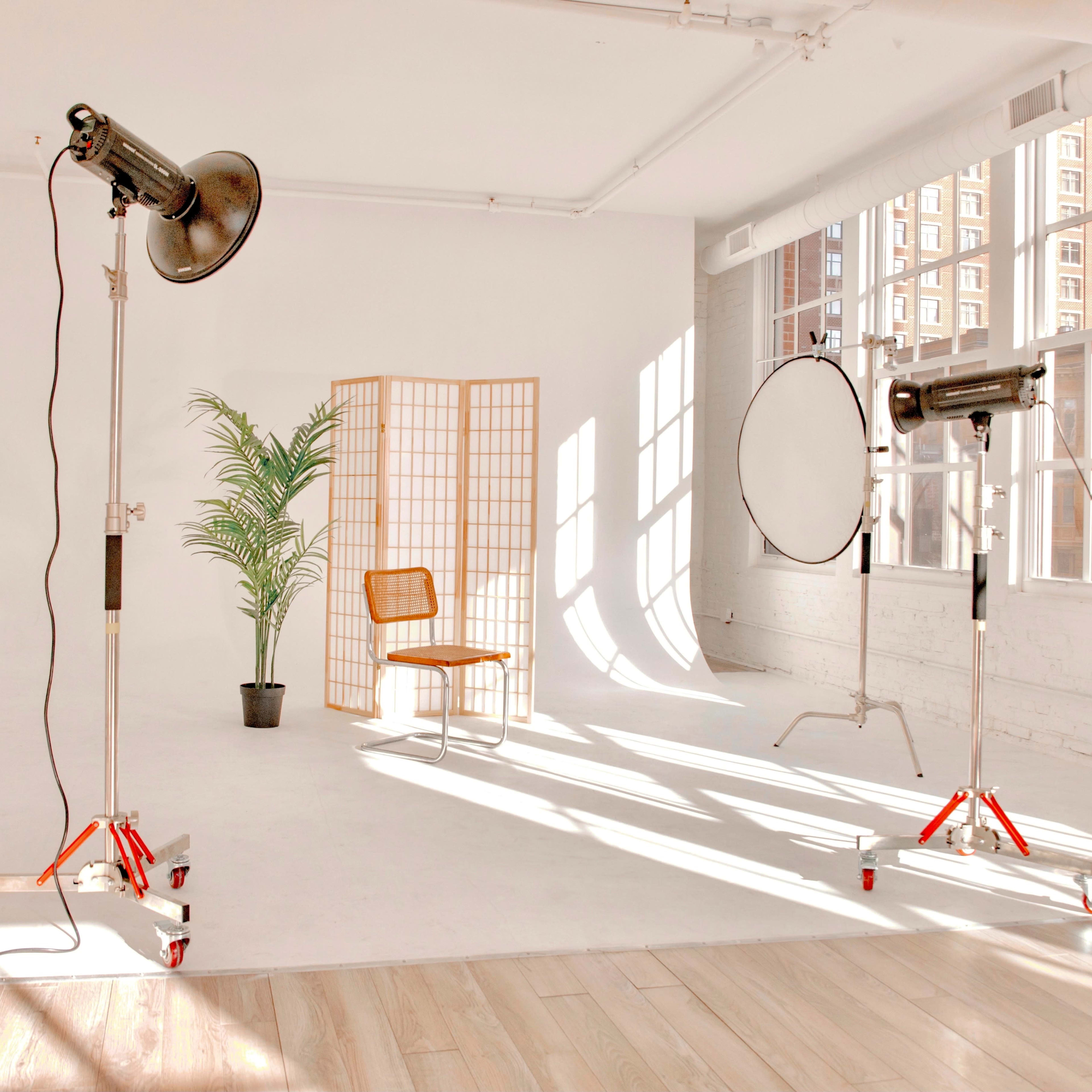A bright studio space with two large lights, a round reflector, a folding screen, a potted plant, and a single wooden chair placed on a wooden floor.
