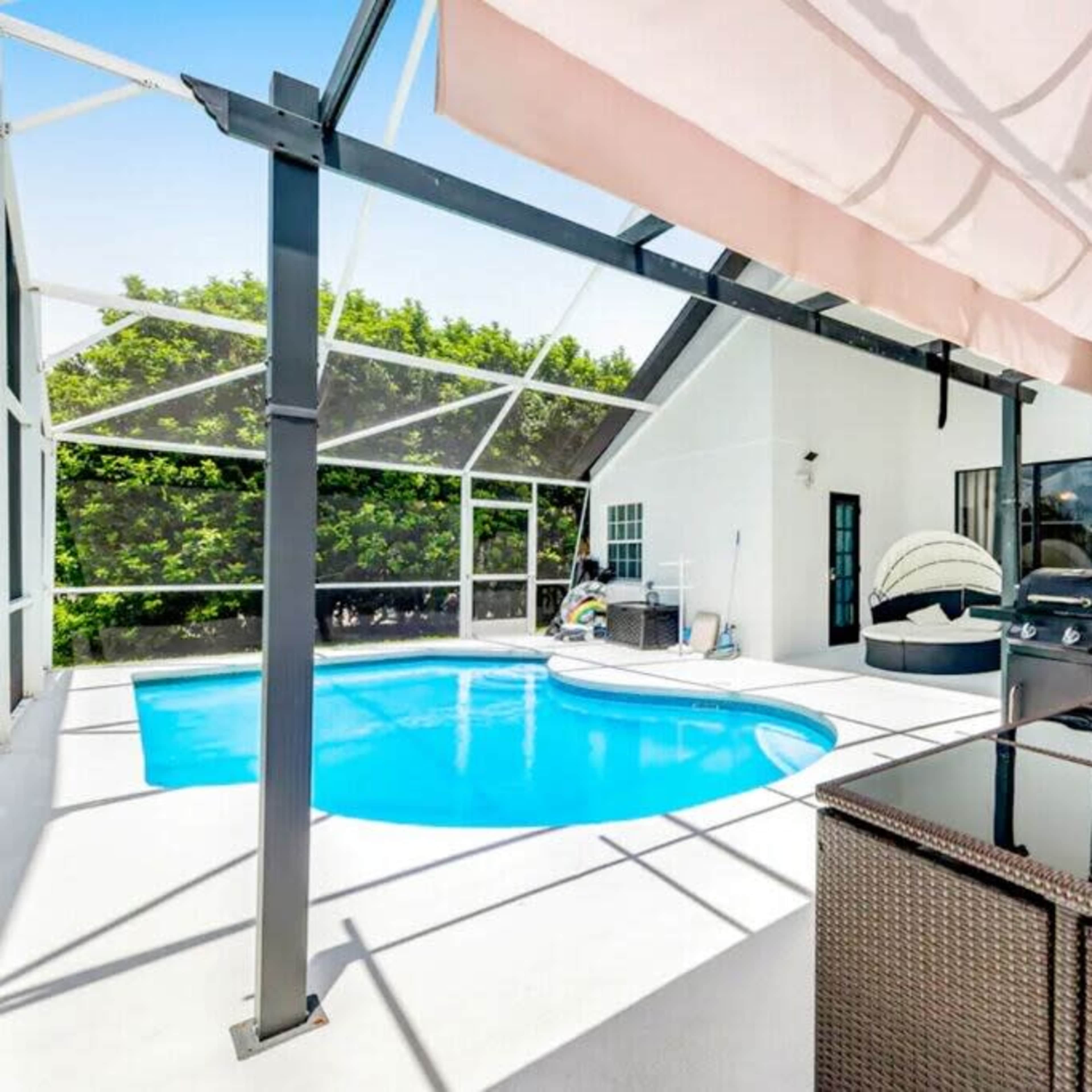 A backyard with a swimming pool, shaded seating area, and a barbecue grill under a covered patio.