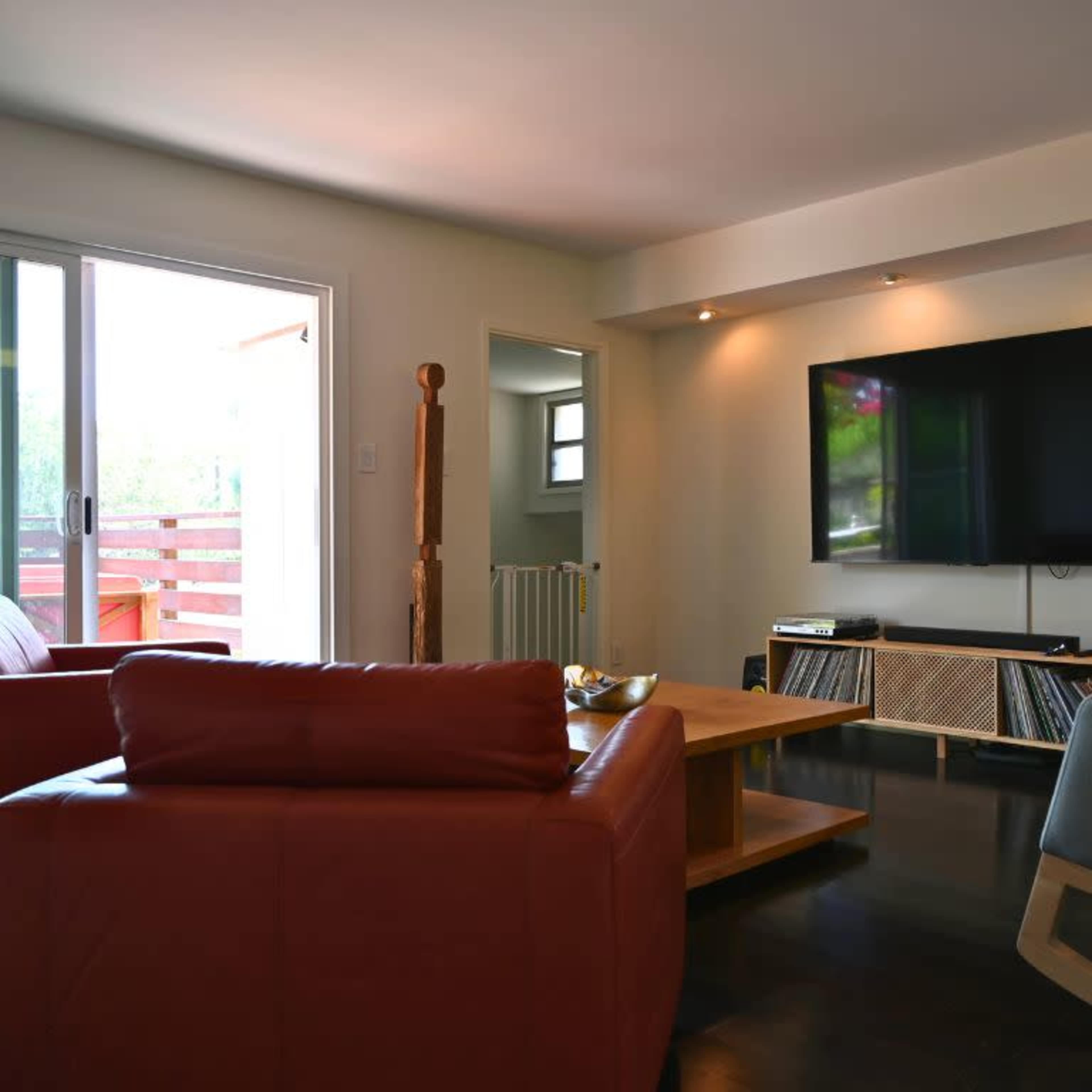 A modern living room features a large flat-screen TV mounted on a wall, with a coffee table, two armchairs, and a sliding glass door leading to an outdoor area.