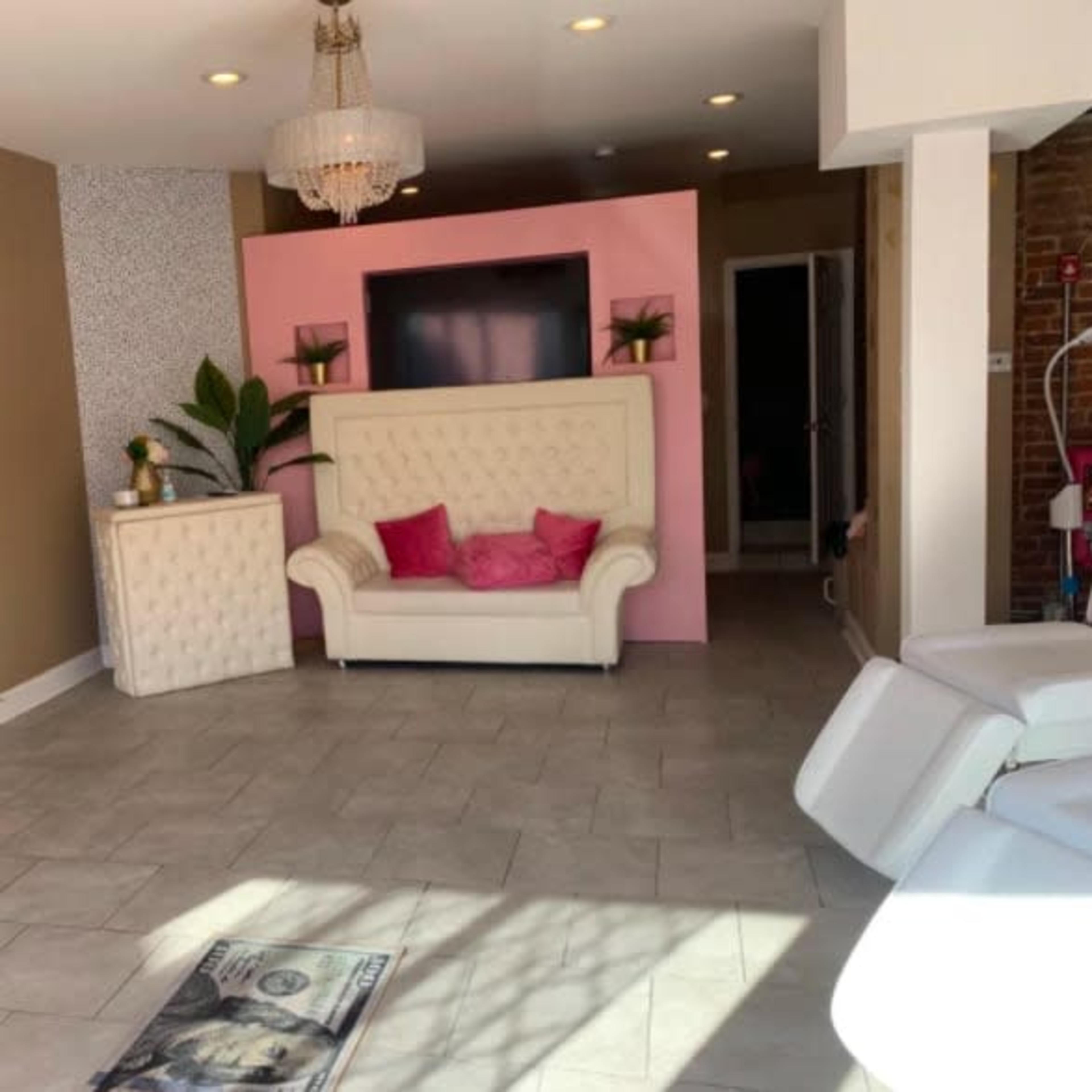 A spacious, brightly lit room with light tile flooring, featuring a white couch with pink accents, a wall-mounted TV, and decorative plants.
