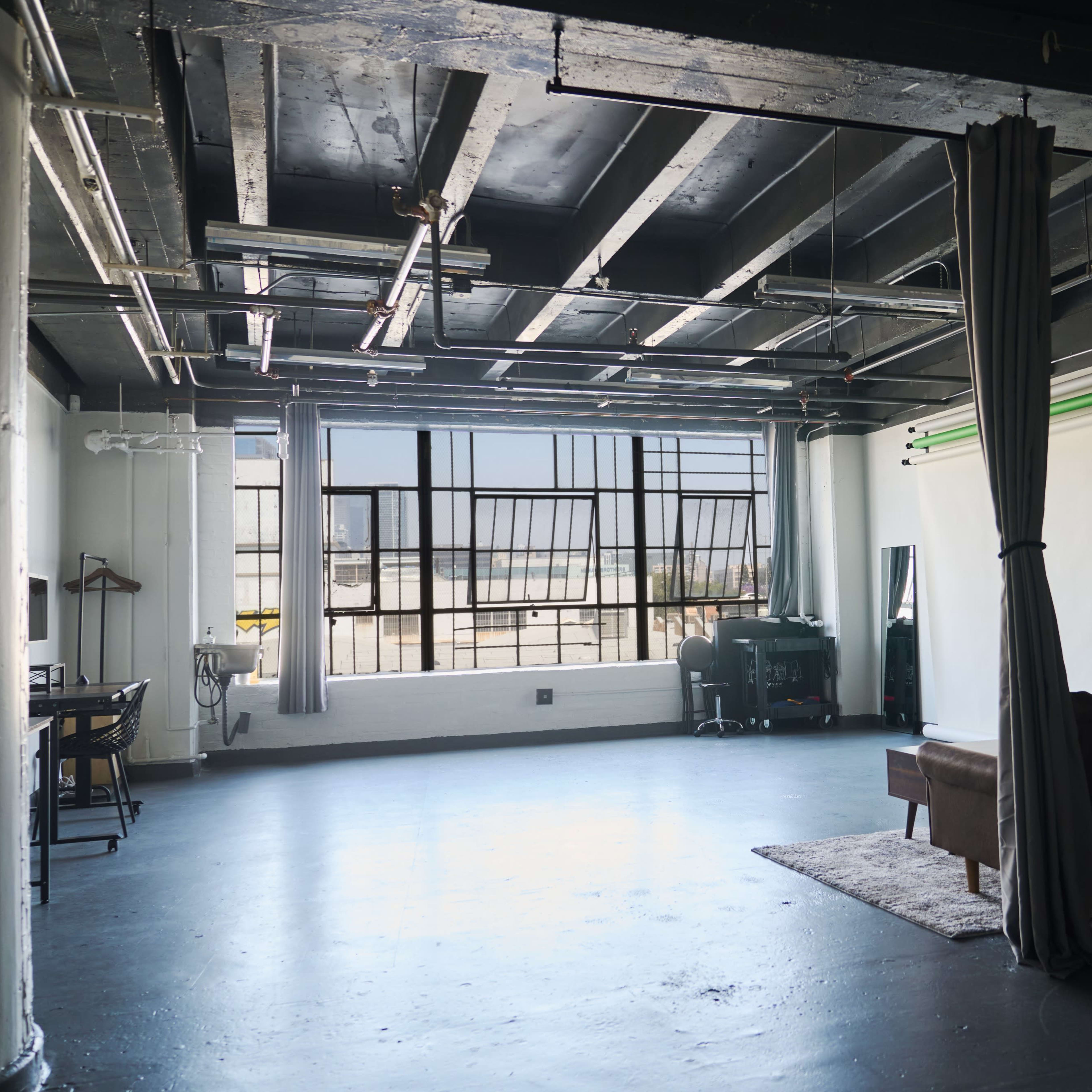 A spacious, industrial-style room with large windows, featuring a couch, a desk setup, and a projector screen against one wall.